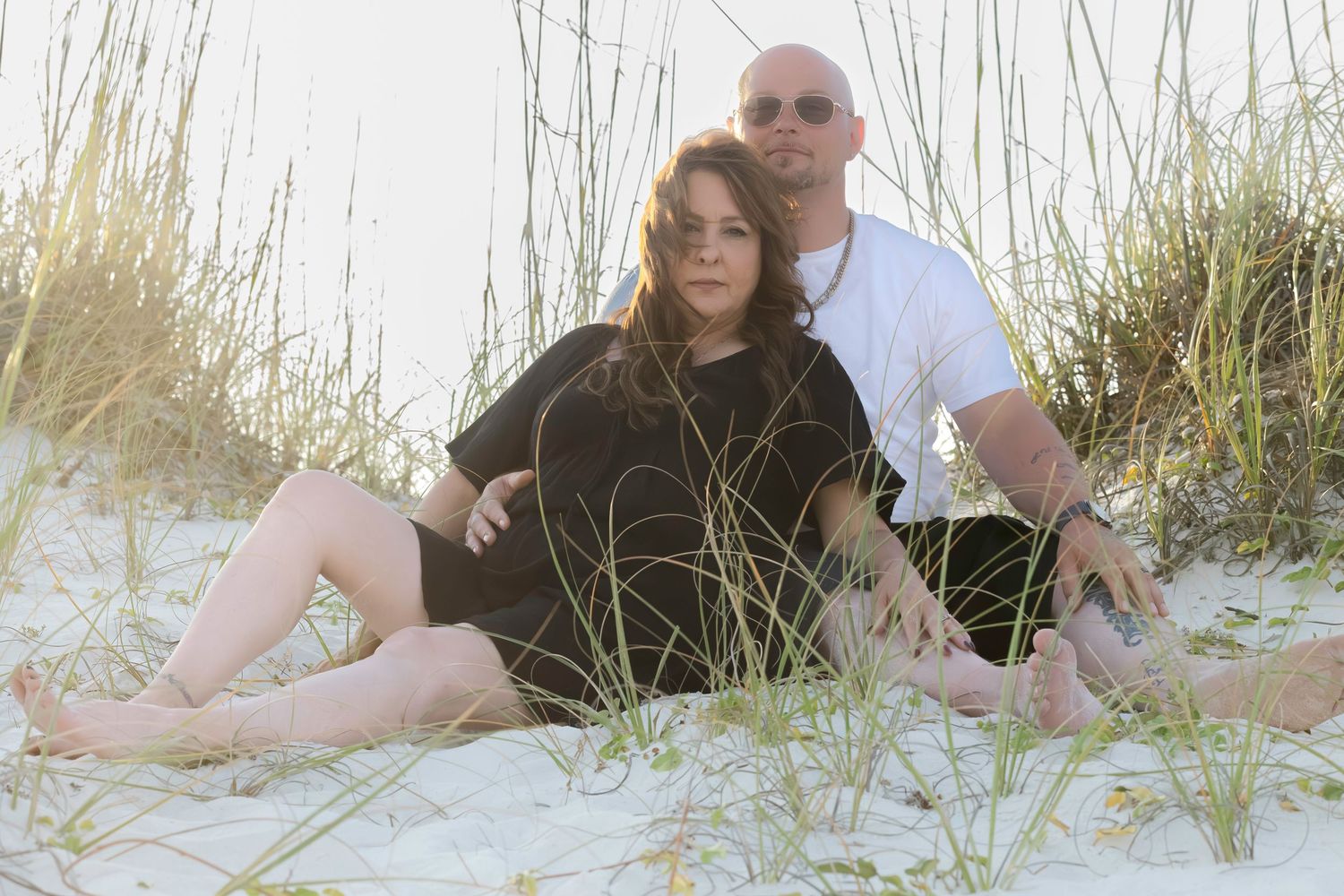 Couple sitting together on white sand beach dunes in casual attire during golden hour.
