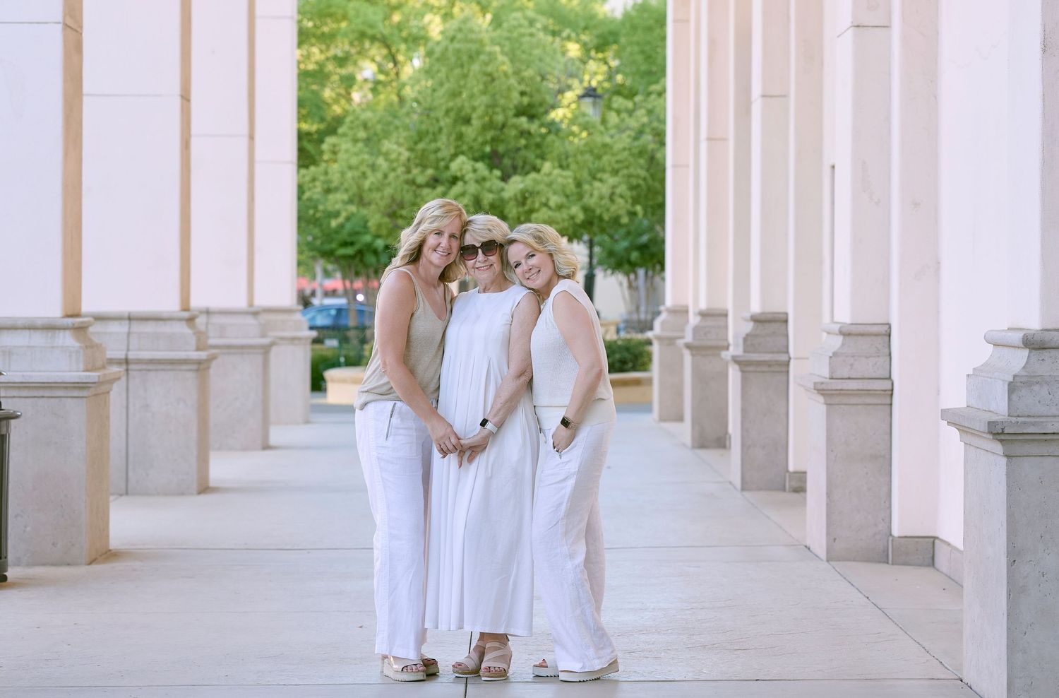 Three people wearing flowing white dresses pose together between elegant stone columns on an outdoor walkway.