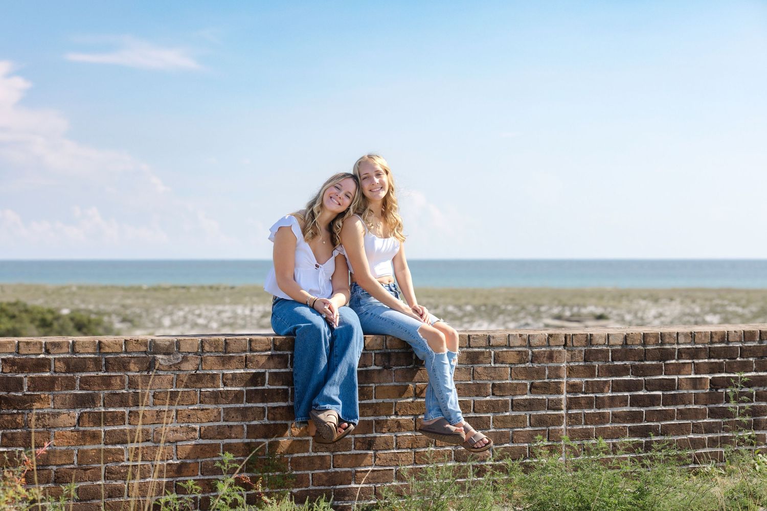 Two people in jeans sit casually on a brick wall overlooking a scenic beach view on a sunny day.