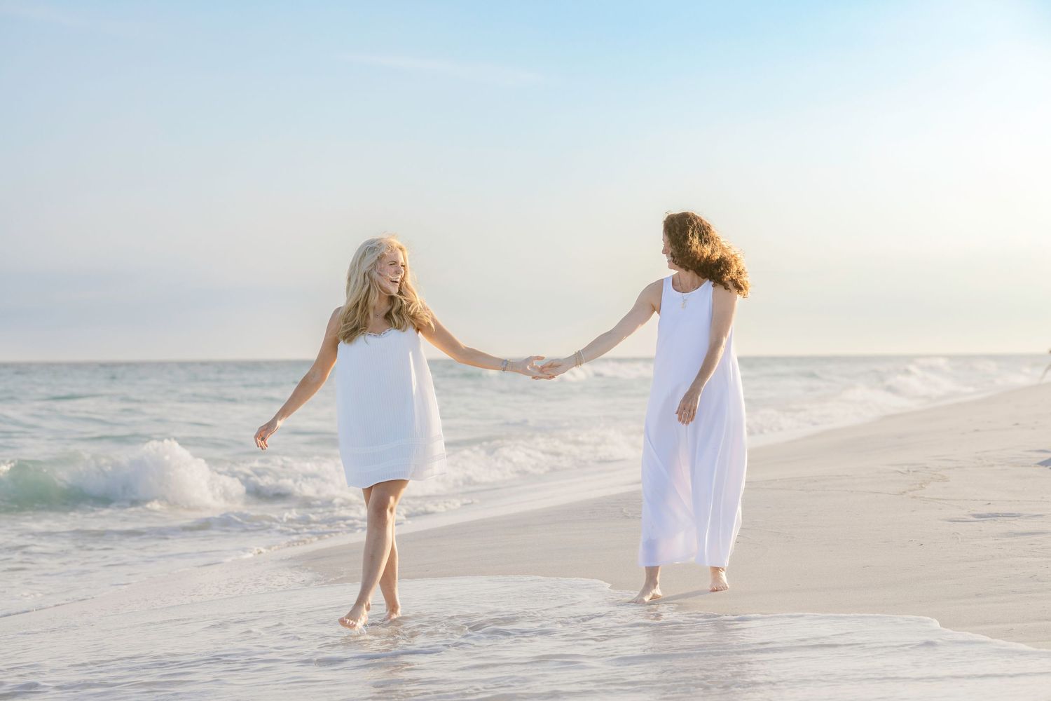 Two girls in white dresses hold hands walking along a sandy beach with ocean waves in the background.