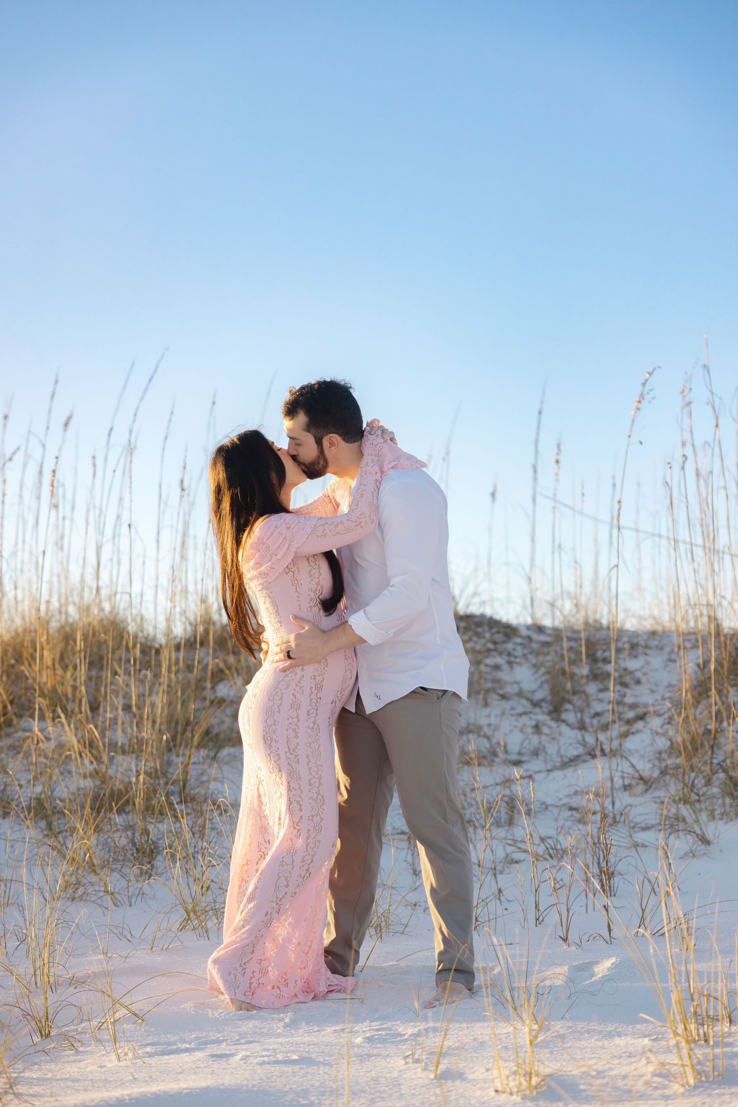 Couple sharing a romantic kiss on white sand dunes with sea oats at golden hour beach setting.