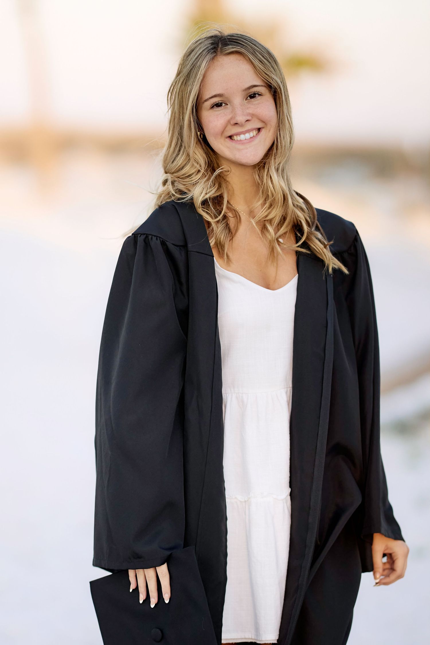 Smiling blonde young woman in black graduation gown and white dress posing outdoors.