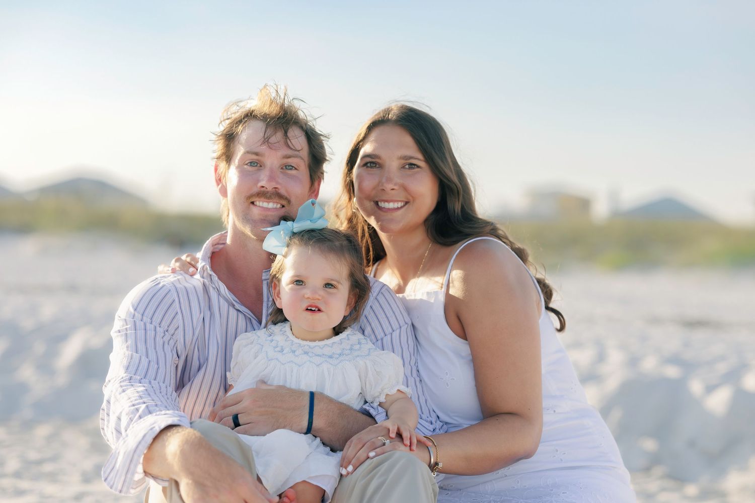 Happy family of three posing on a sandy beach, parents smiling with baby in white outfit.