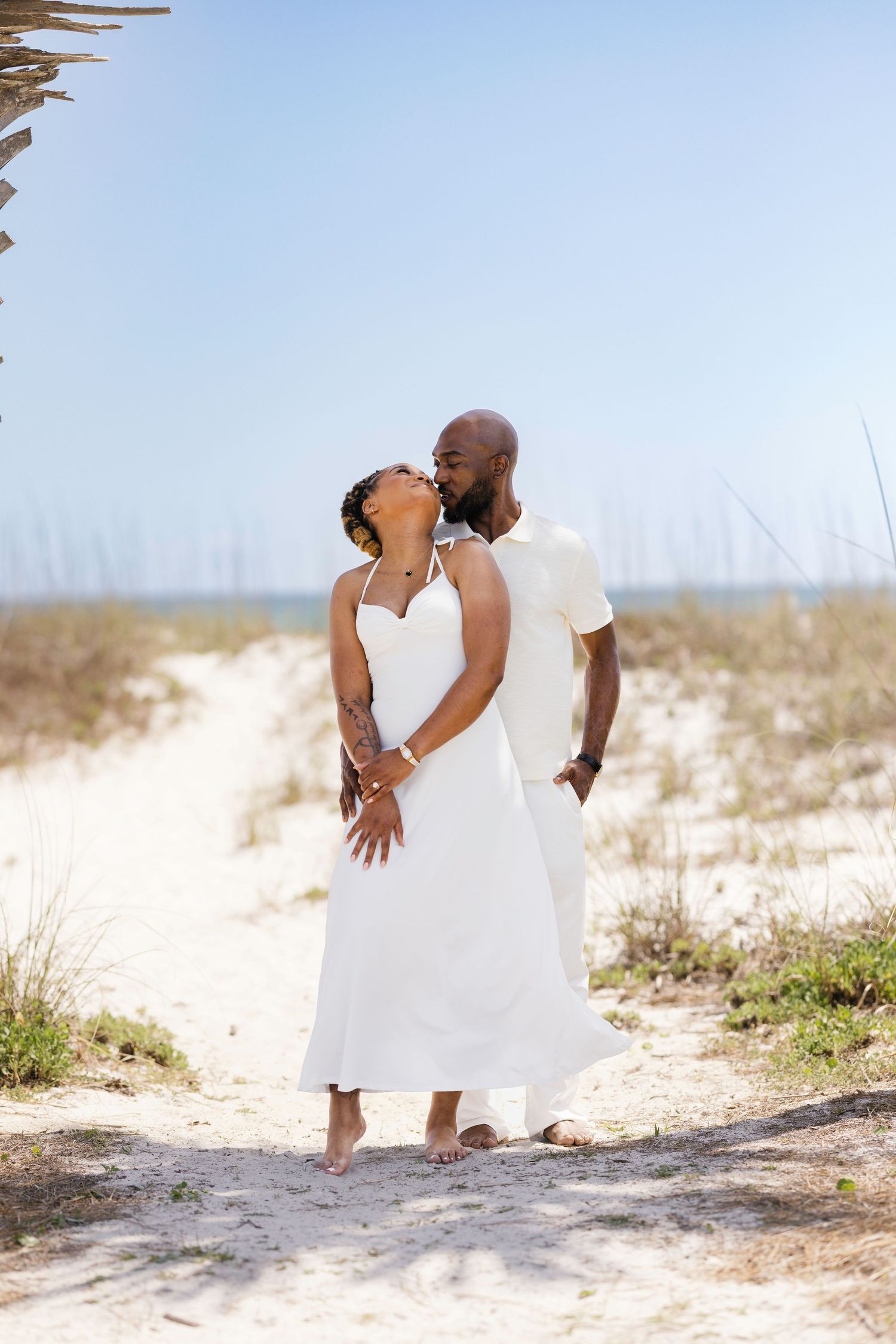 A couple shares a kiss on a sandy beach path, both dressed in white on a sunny day.