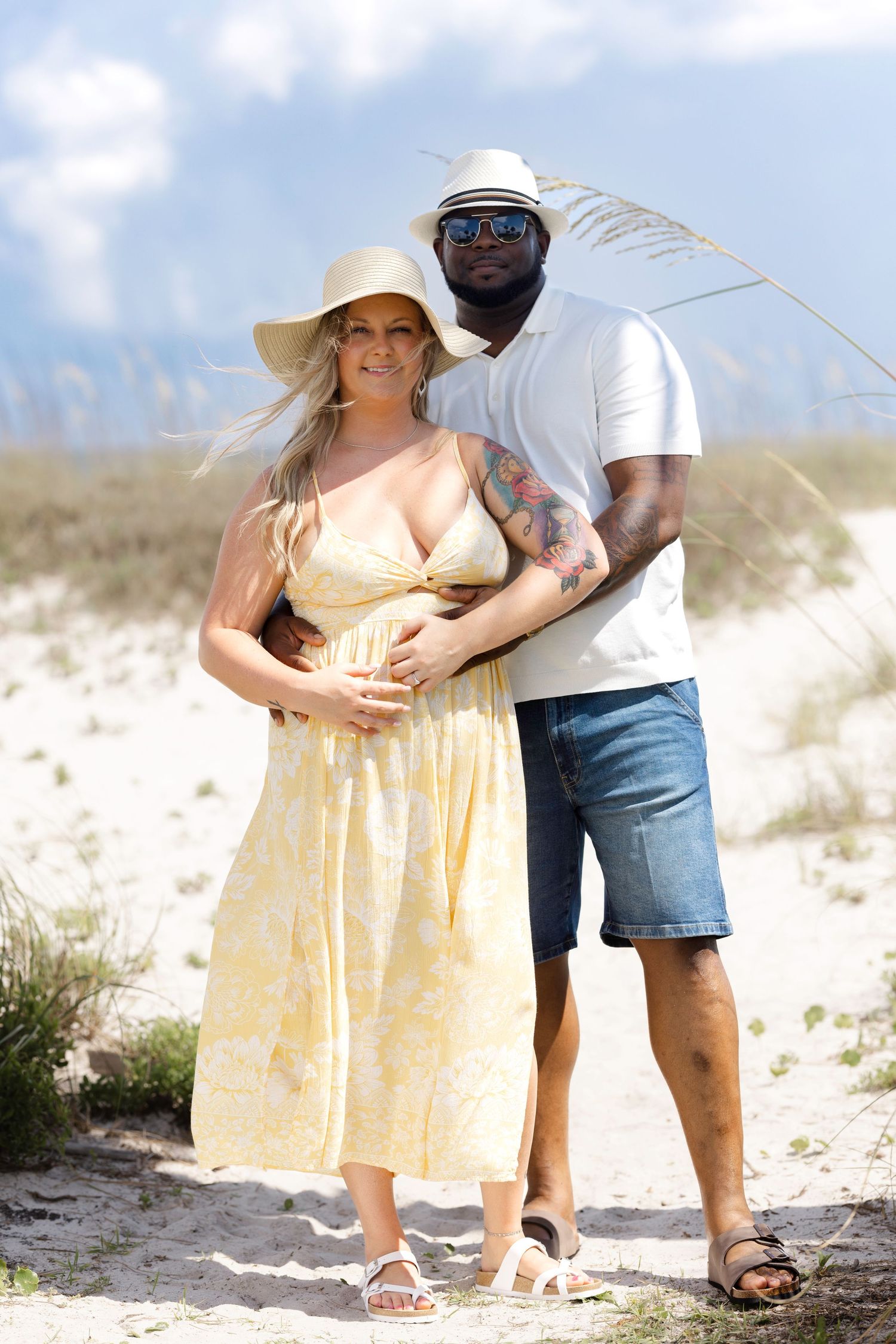Pregnant woman in yellow dress and sun hat poses with man in white shirt and denim shorts on sandy beach.