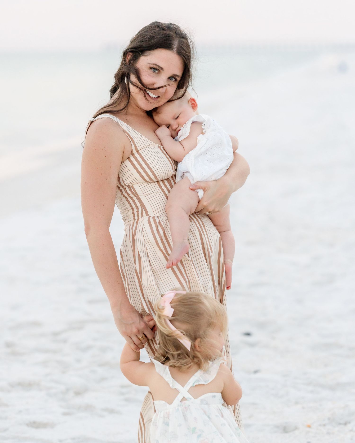 Beautiful brunette mother in striped dress holds newborn while toddler daughter hugs her at beach.