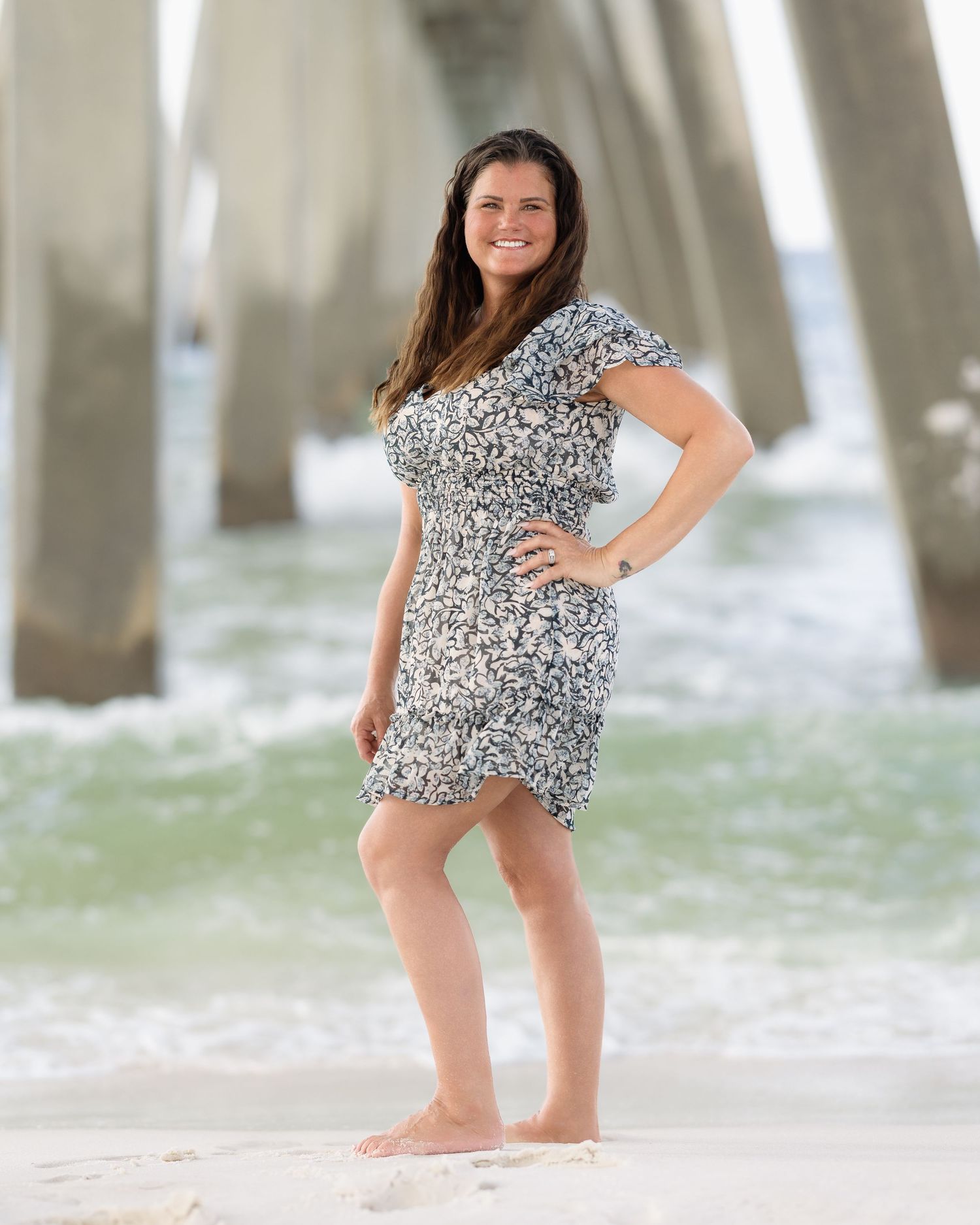 Woman in floral dress posing barefoot on beach under pier with ocean waves behind her.