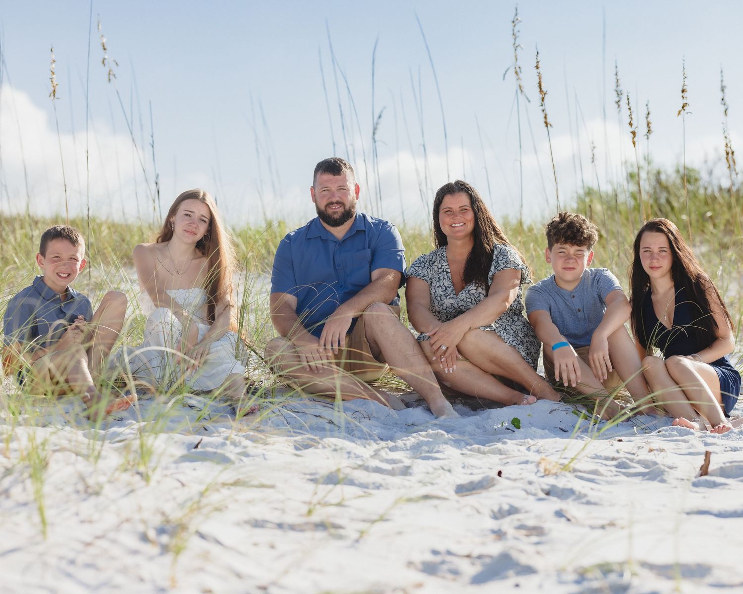 Family of six posing together on a white sand beach with sea oats and blue sky backdrop.