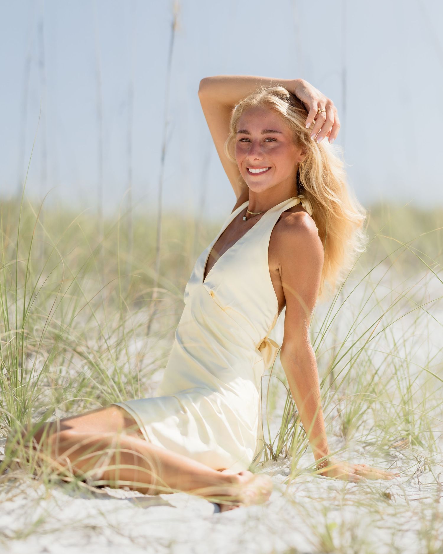 Smiling blonde woman in white dress sitting on sandy beach dunes with sea oats on a sunny day.