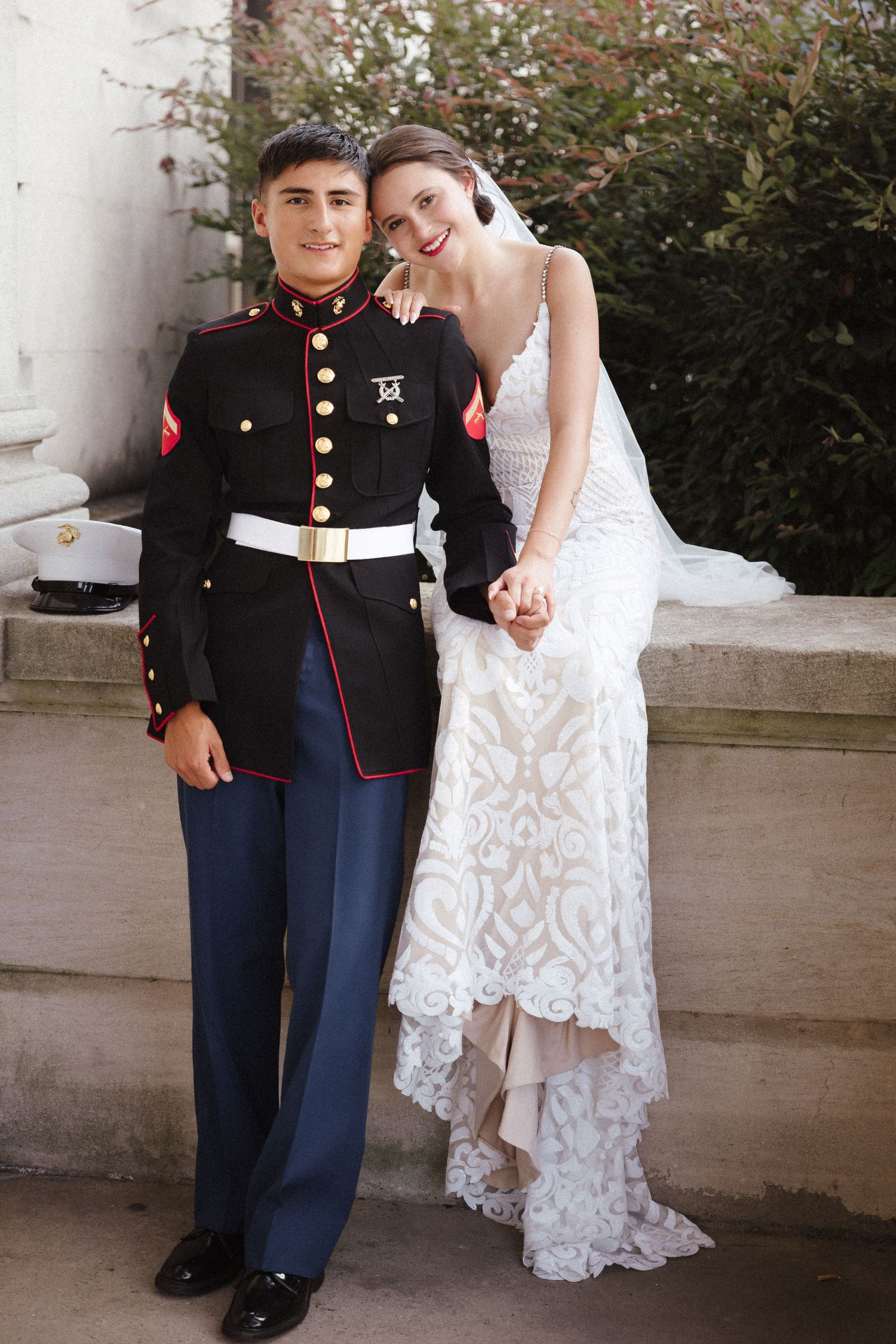 Marine in dress uniform stands with bride in white lace gown, posing together outdoors near stone columns.