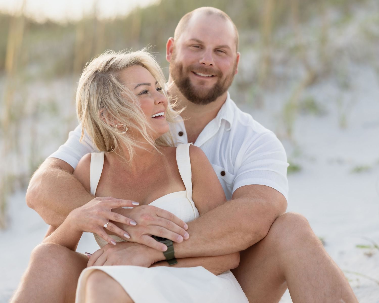 Happy couple sitting together on a sandy beach, woman in white dress laughing, man in white shirt smiling warmly.