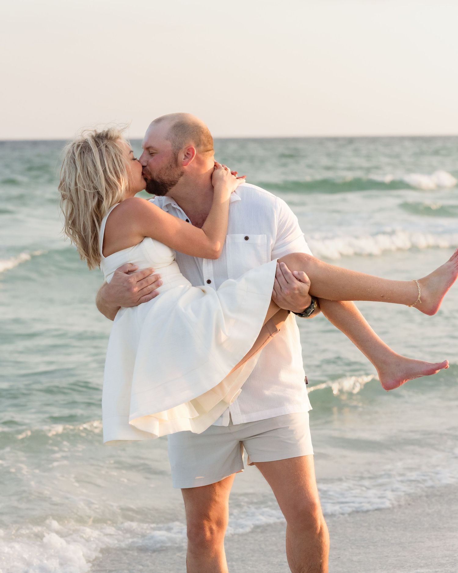 Man in white shirt lifting and kissing woman in white dress on a sunny beach.