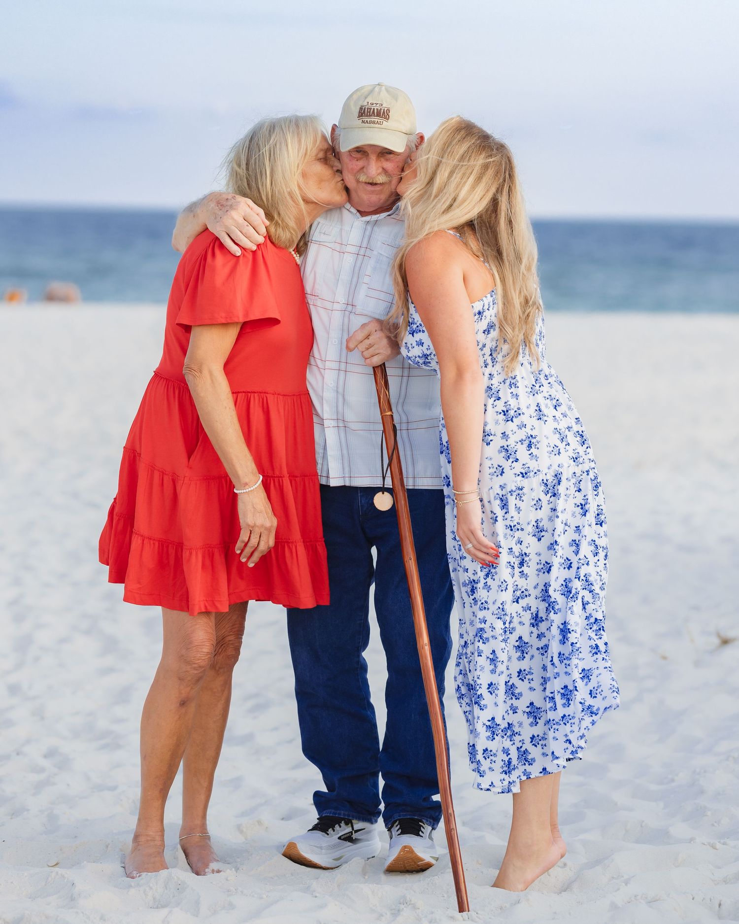Elderly man with cane receives kisses from two women in blue and red dresses on a sunny beach.