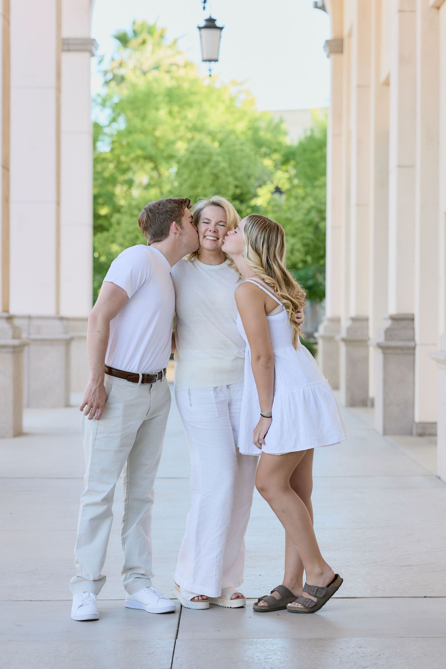 Three people in white summer outfits share a tender moment together in a columned outdoor walkway.