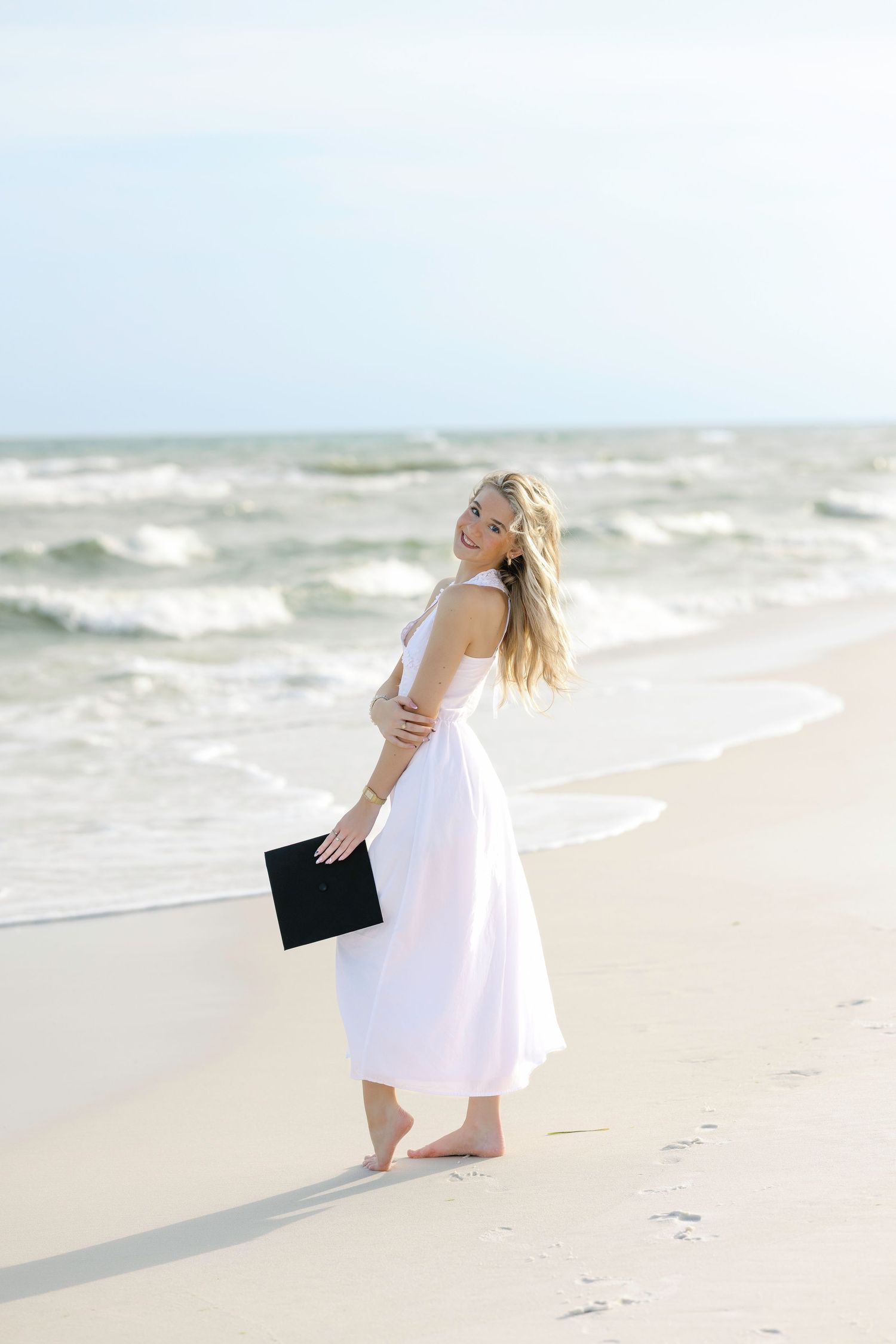 Seaside photoshoot with waves and beach backdrop featuring flowing white dress and dark clutch.