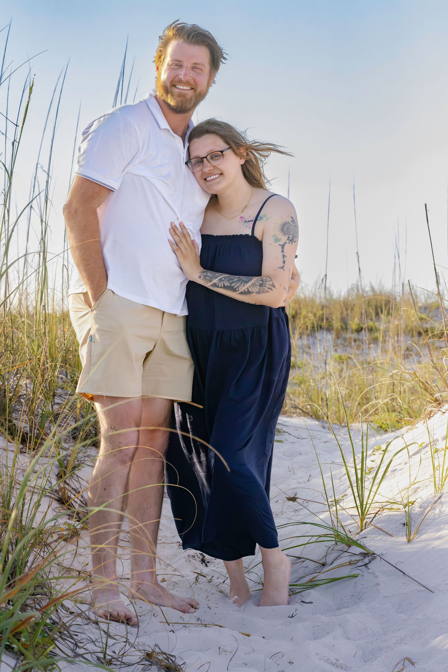 Couple embracing on sandy beach dunes, woman in navy dress with tattoos, man in white polo and khaki shorts, smiling.