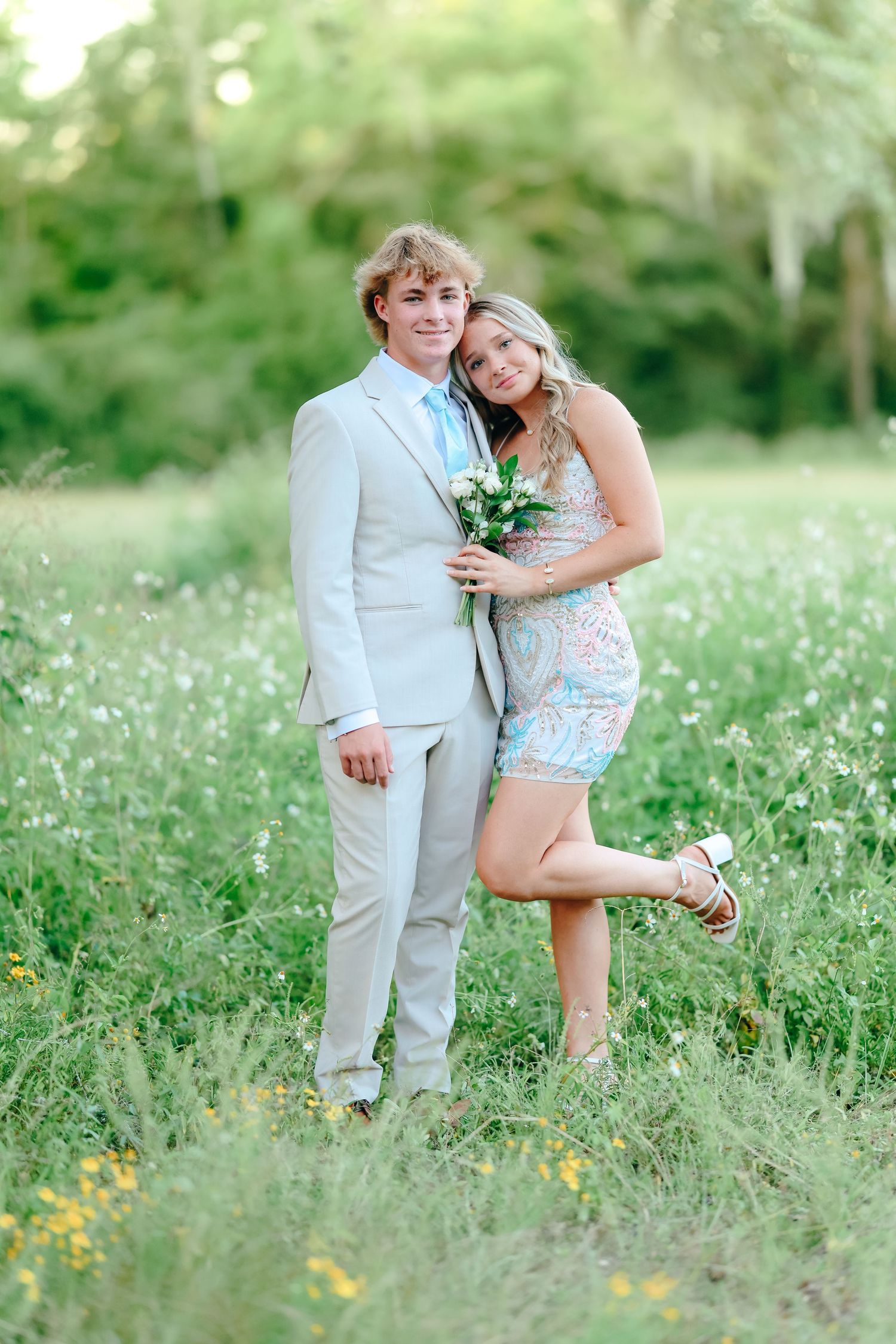 A young couple in prom attire posing in a field of wildflowers, girl in blue dress leaning on suited boy.