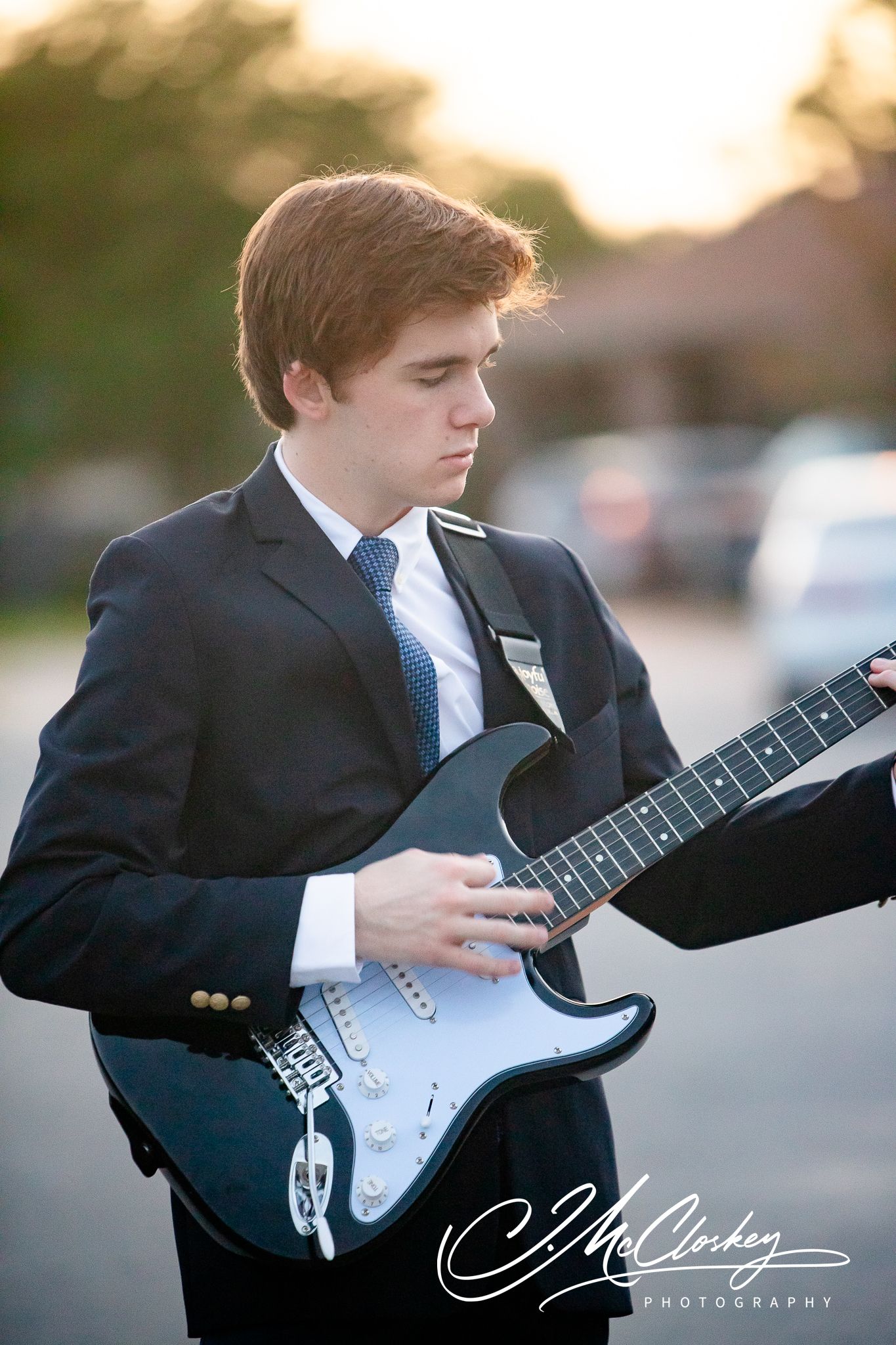 A musician in formal attire plays an electric Stratocaster guitar outdoors at sunset with soft backlighting.
