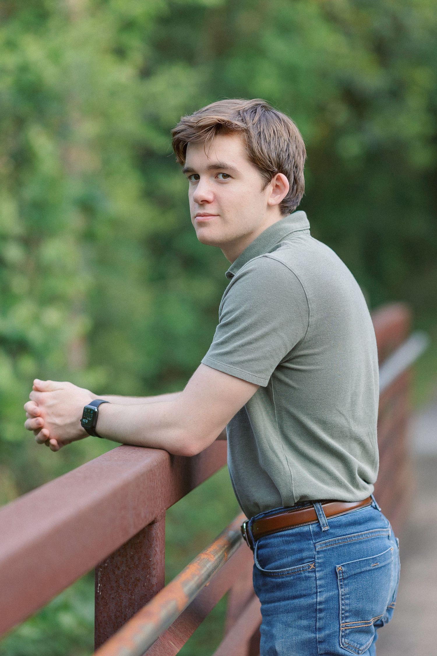 Young man in green polo and jeans leaning on rustic bridge railing outdoors with lush green trees in background.