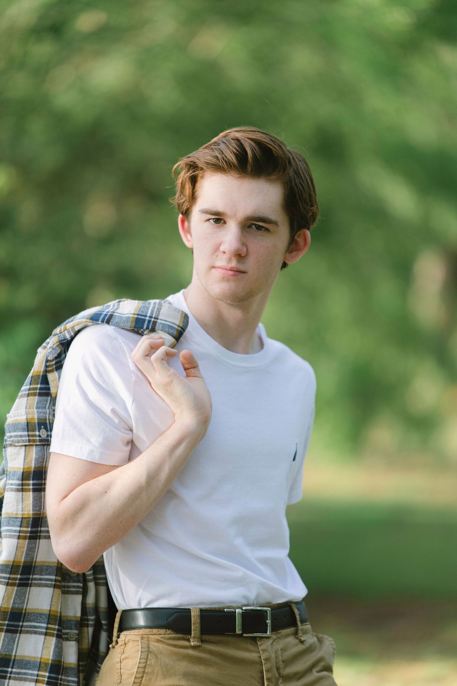 Young man in white t-shirt and plaid jacket poses outdoors in a lush green park setting.