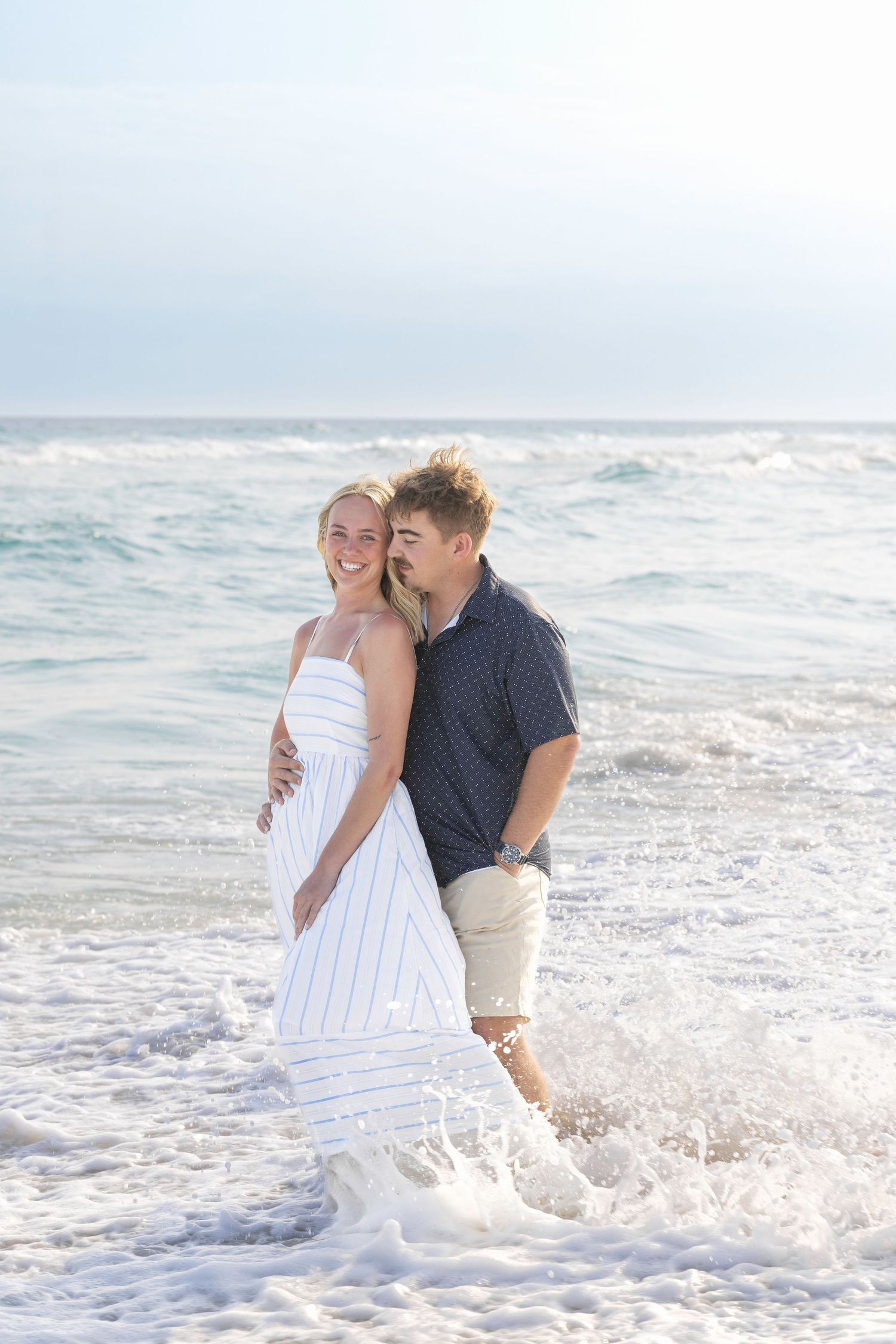 A couple stands in ocean waves on a sunny beach, the woman in a white dress smiling as the man embraces her from behind.