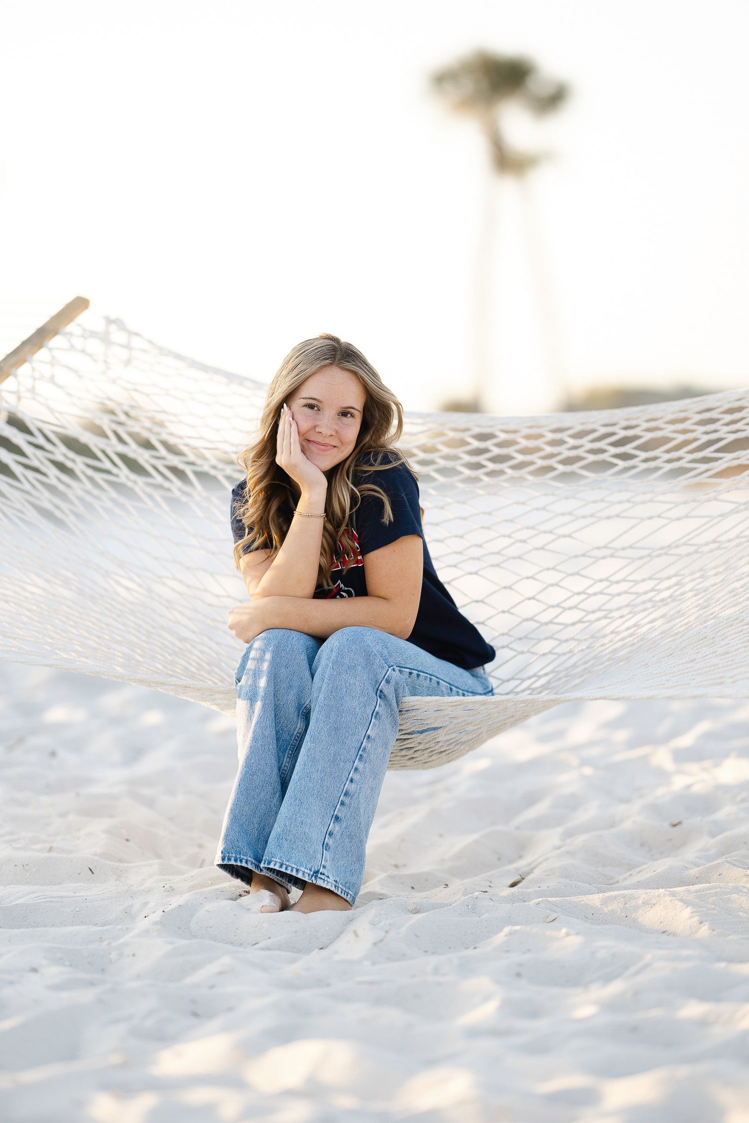 Person relaxes in white hammock on sandy beach wearing blue jeans and navy top in sunset lighting.