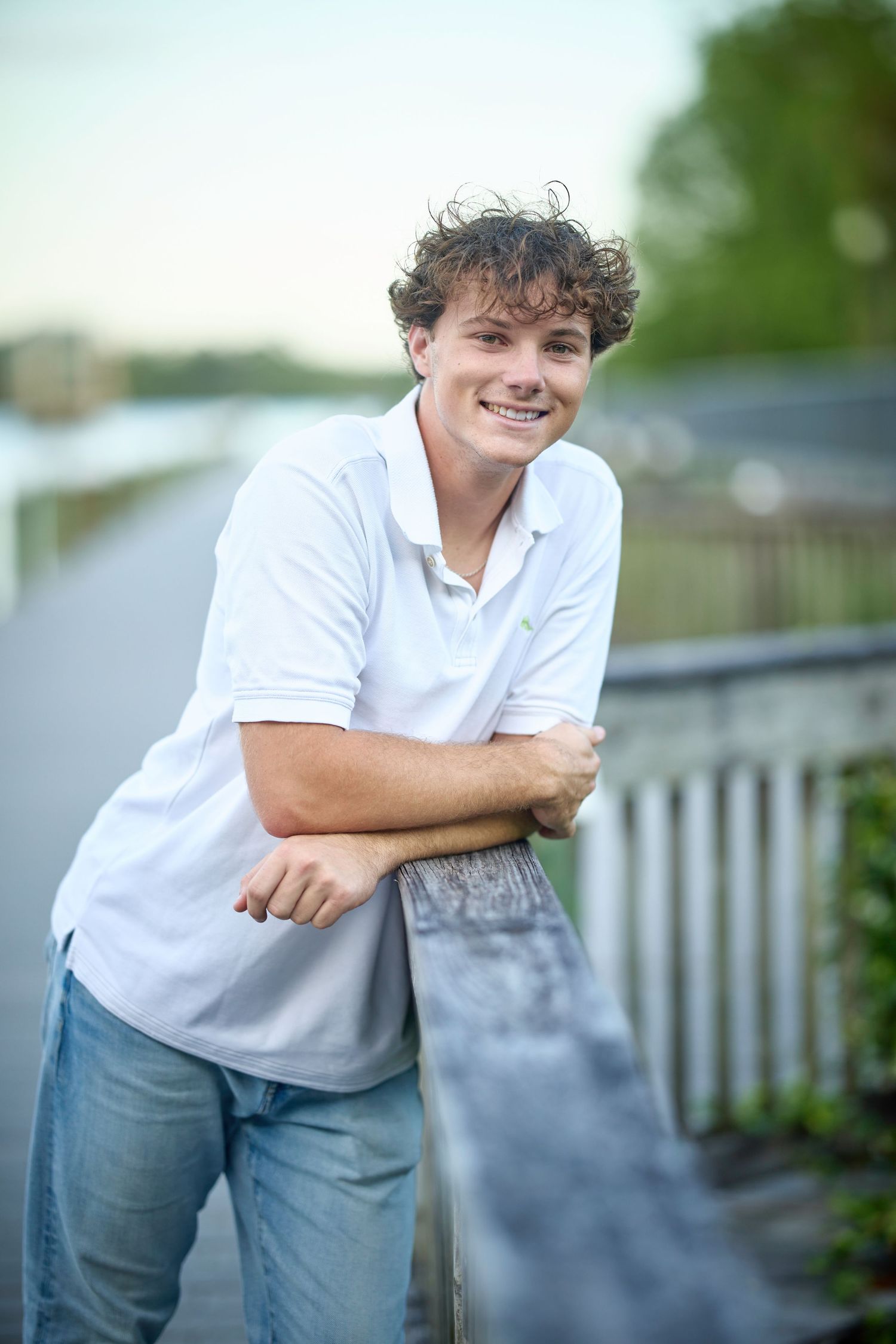 Smiling teenage boy in white polo shirt leaning on railing outdoors near waterway on a bright day.