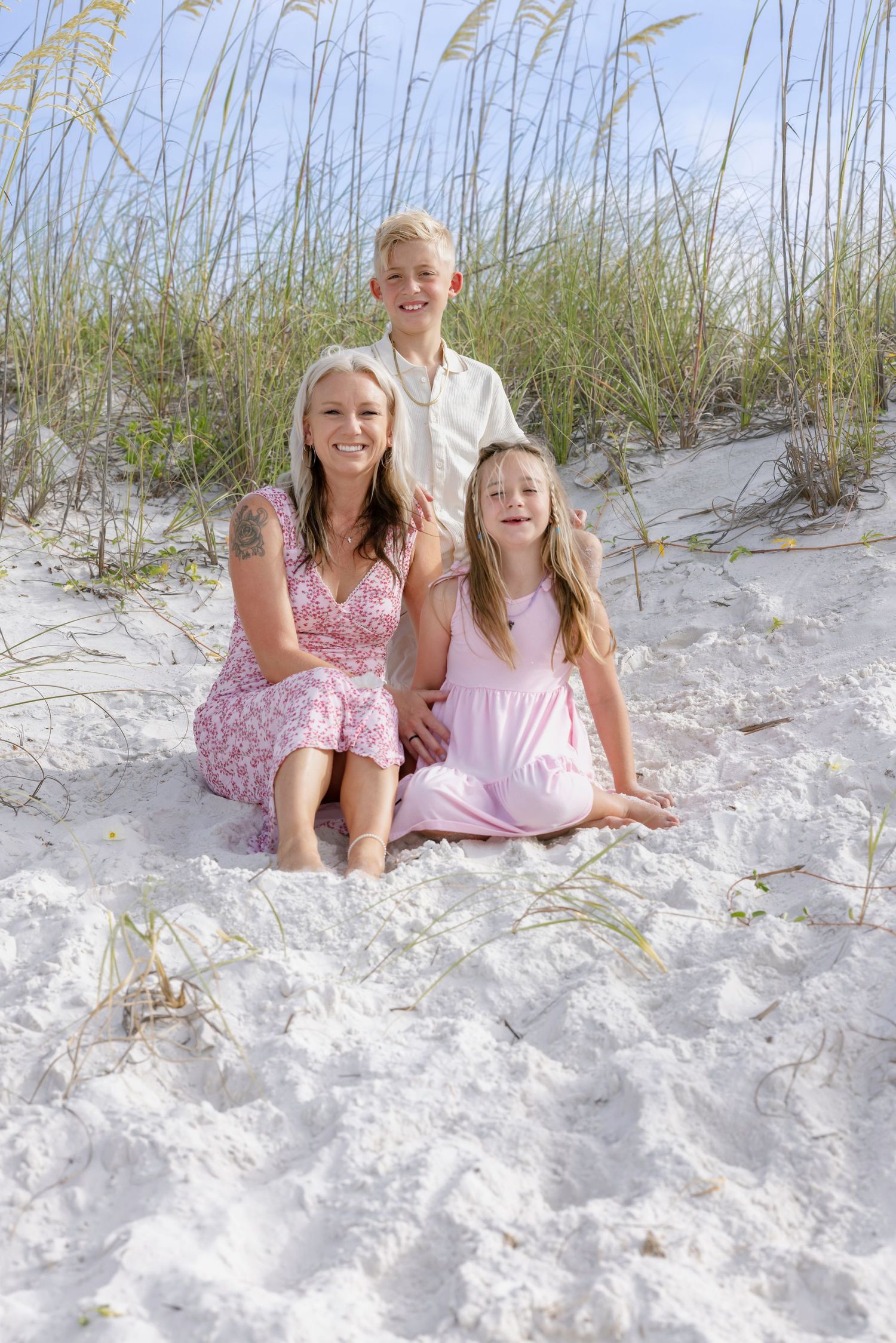 A smiling woman and two children sit together on white sand beach dunes with sea oats in the background.