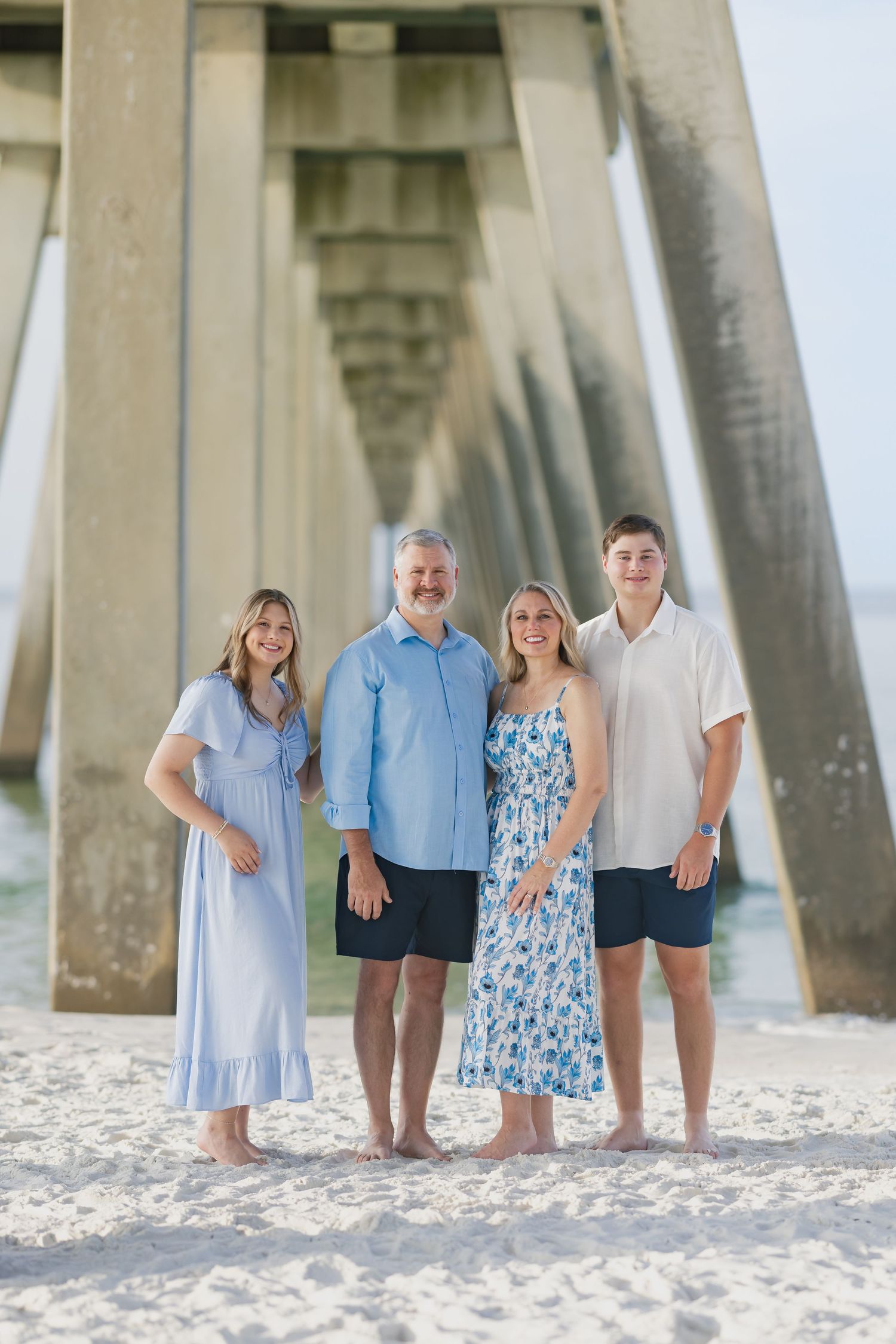 Family of four poses barefoot on white sand beach under a pier, wearing coordinated blue and white outfits.