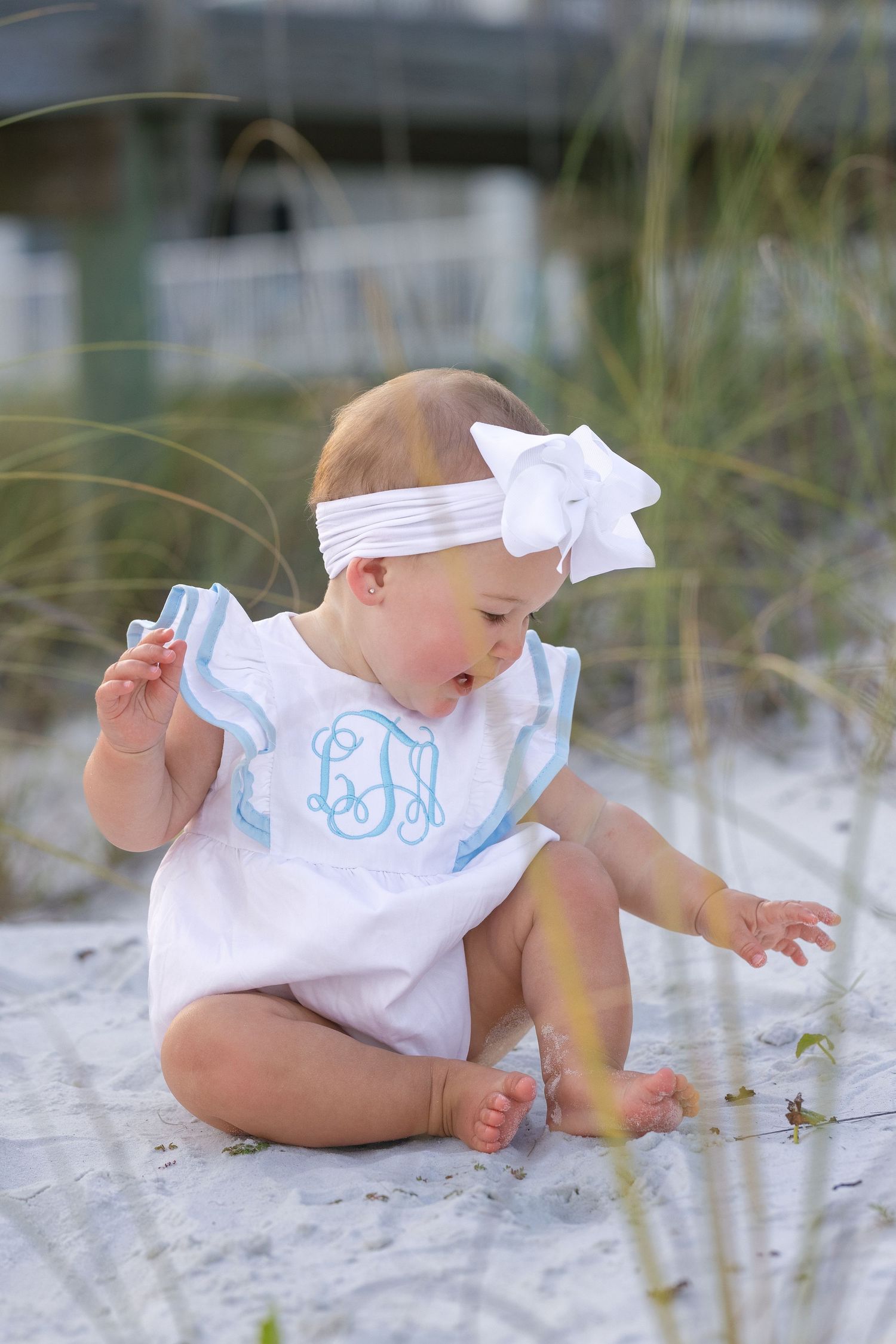 Baby girl in white monogrammed romper and bow headband sitting on white sand beach.