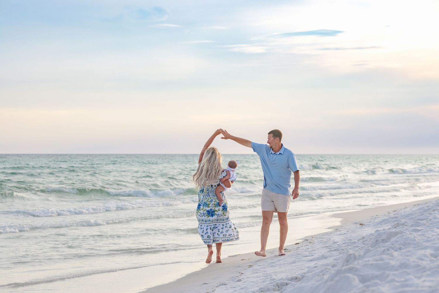 Family dancing on a white sand beach at sunset, parents spinning with baby in arms near ocean waves.