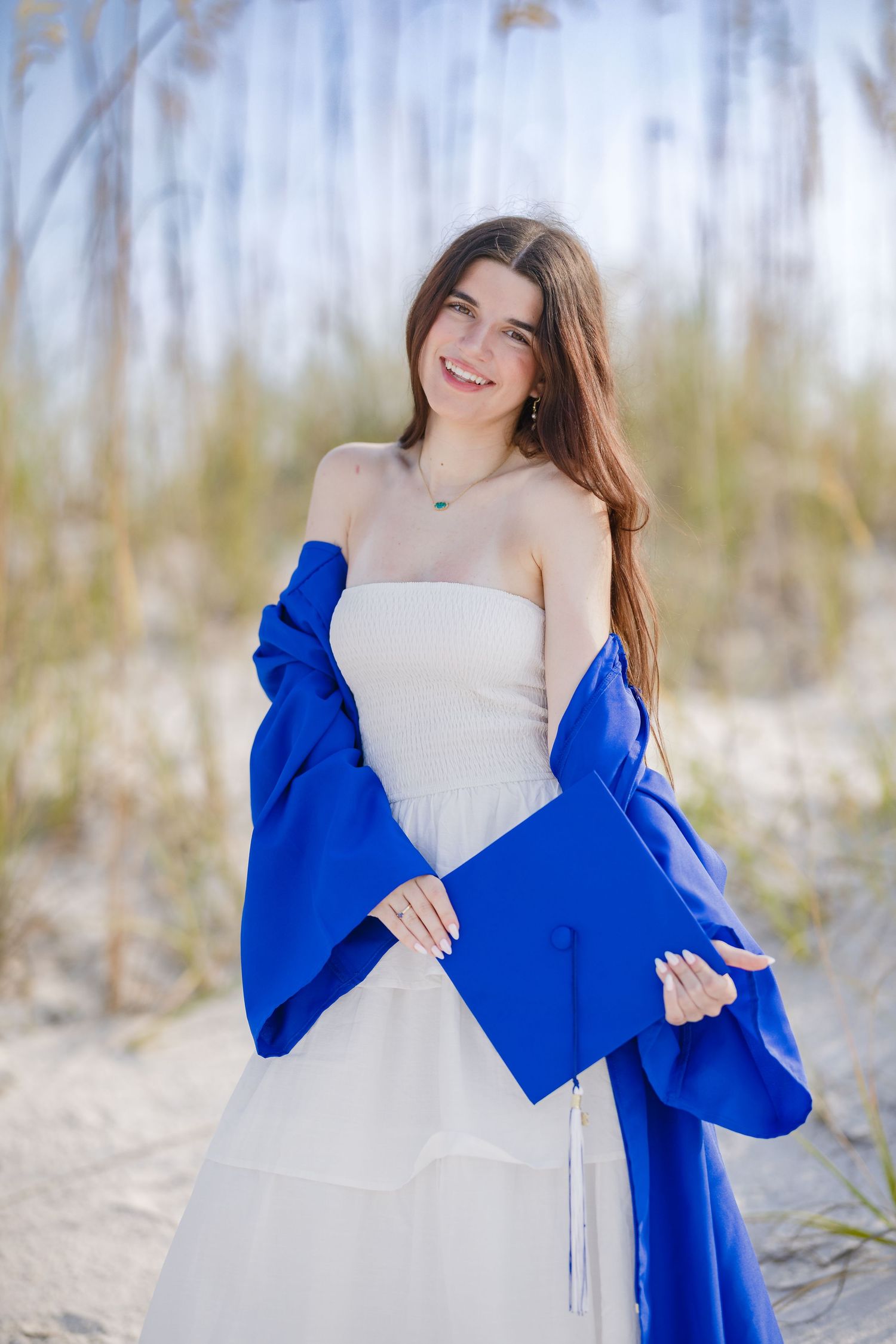 Smiling young woman in white dress holding blue graduation cap and gown on sandy beach.