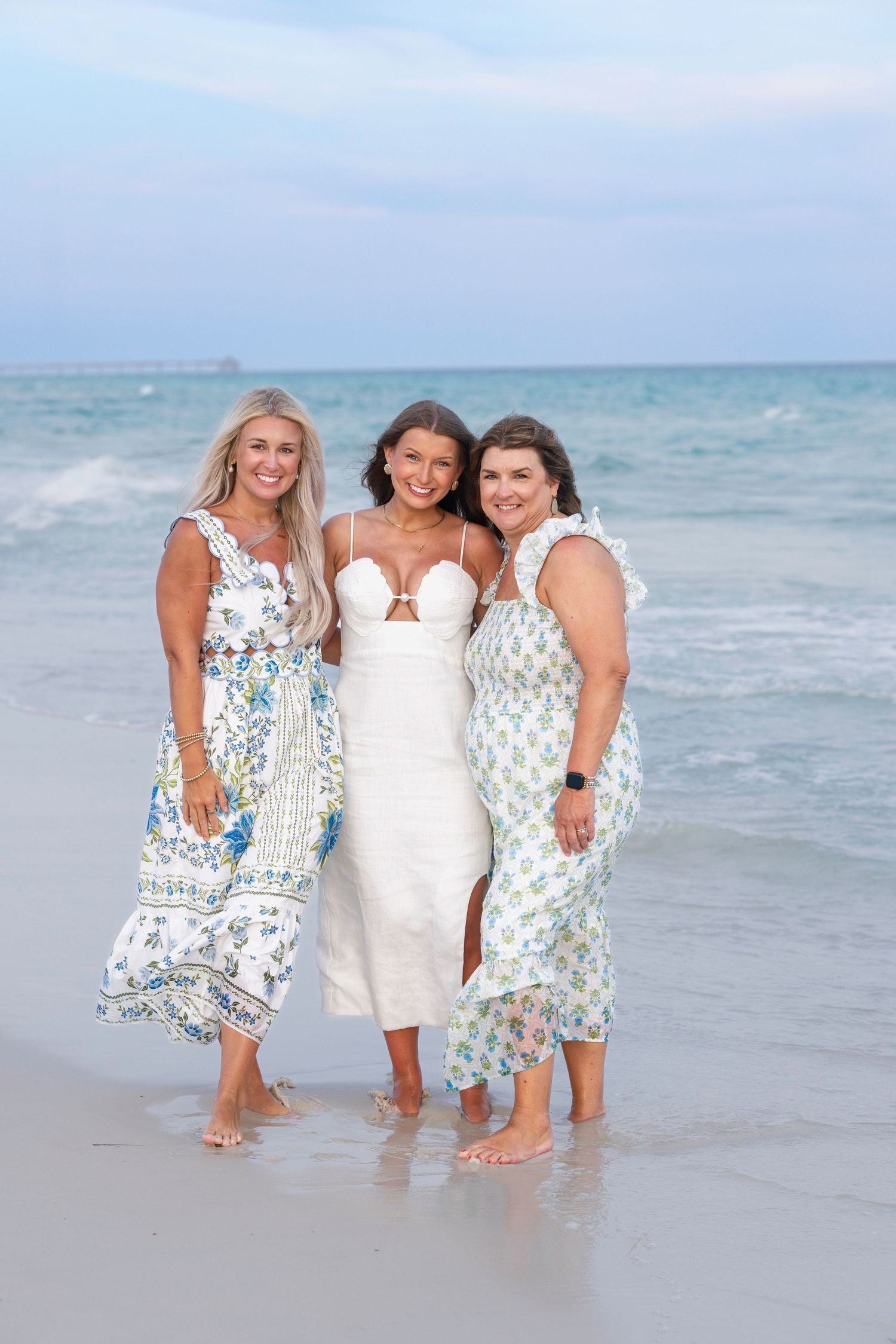 Three women smiling together on a beach, wearing floral and white dresses with ocean waves behind them.