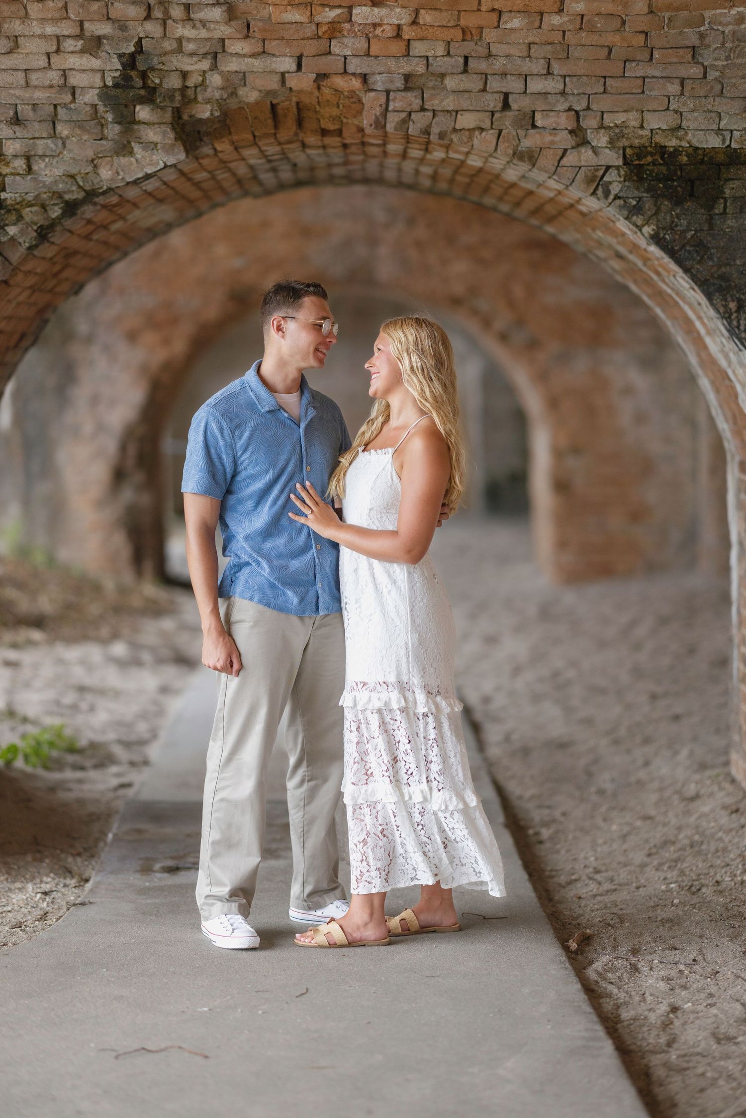 Couple in love standing under brick arches, man in blue shirt and woman in white lace dress, gazing at each other.