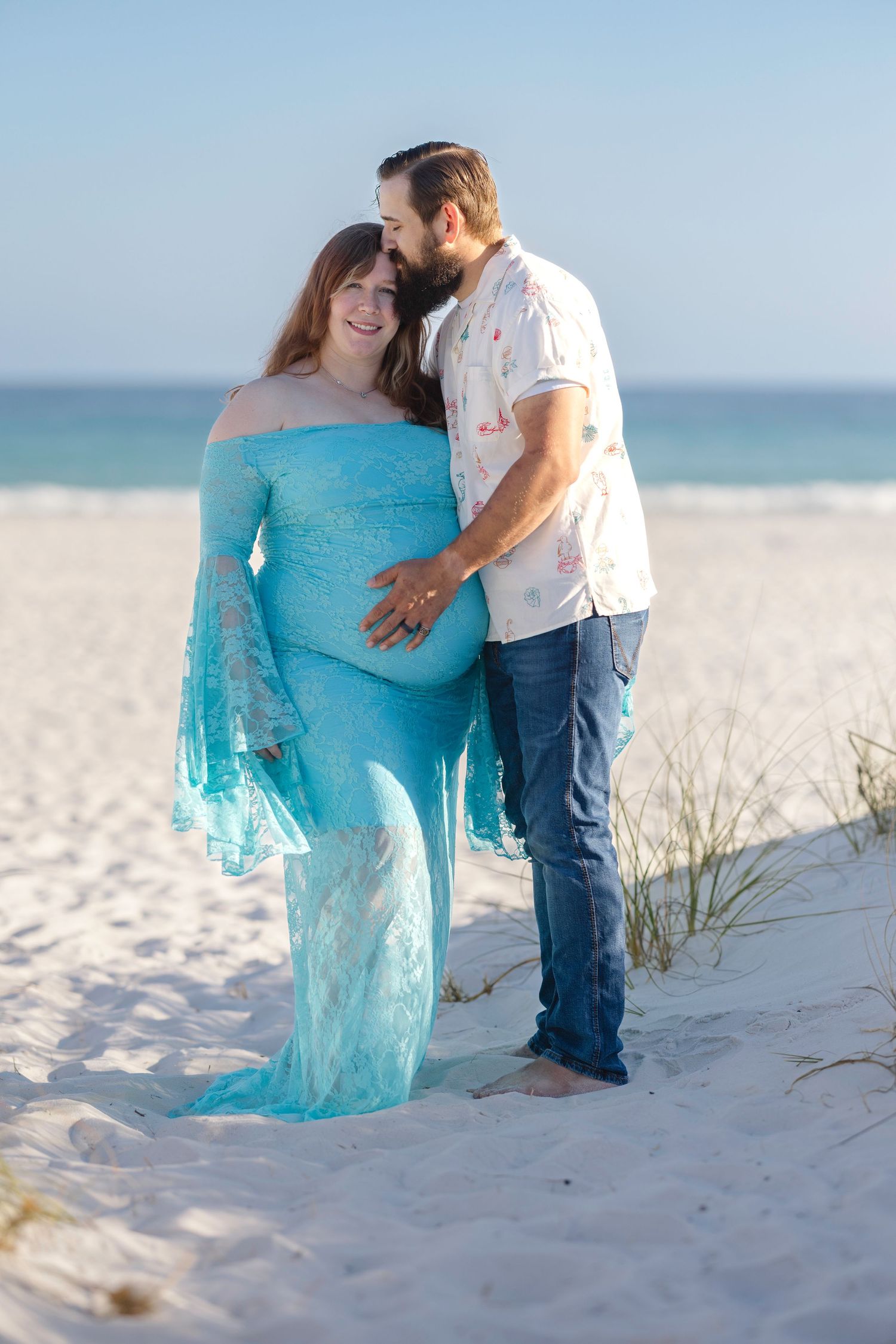 Pregnant woman in teal lace gown embraced by bearded man on white sand beach.
