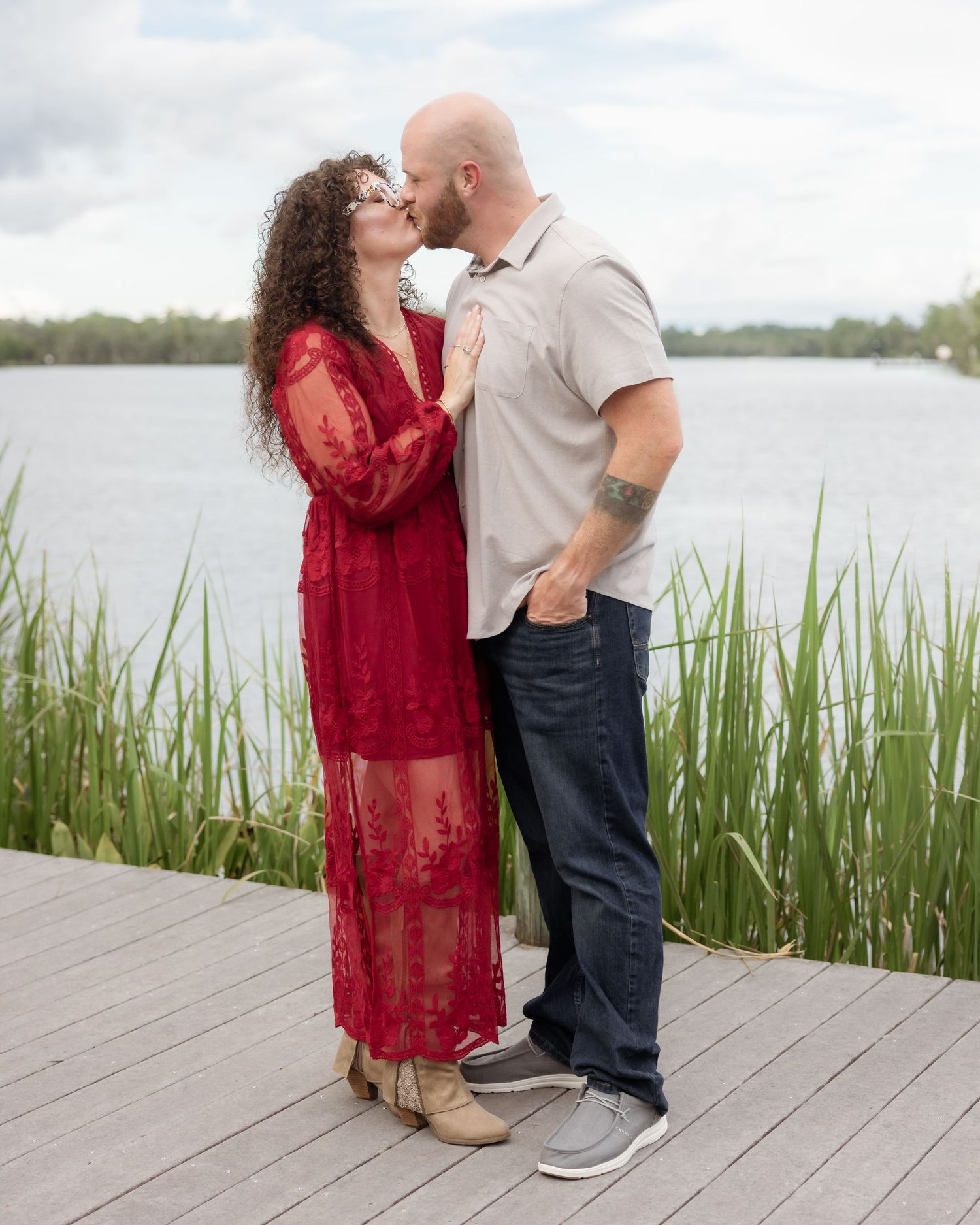 Couple sharing a romantic kiss on a wooden dock by a serene lake, woman in red lace dress, man in gray shirt.
