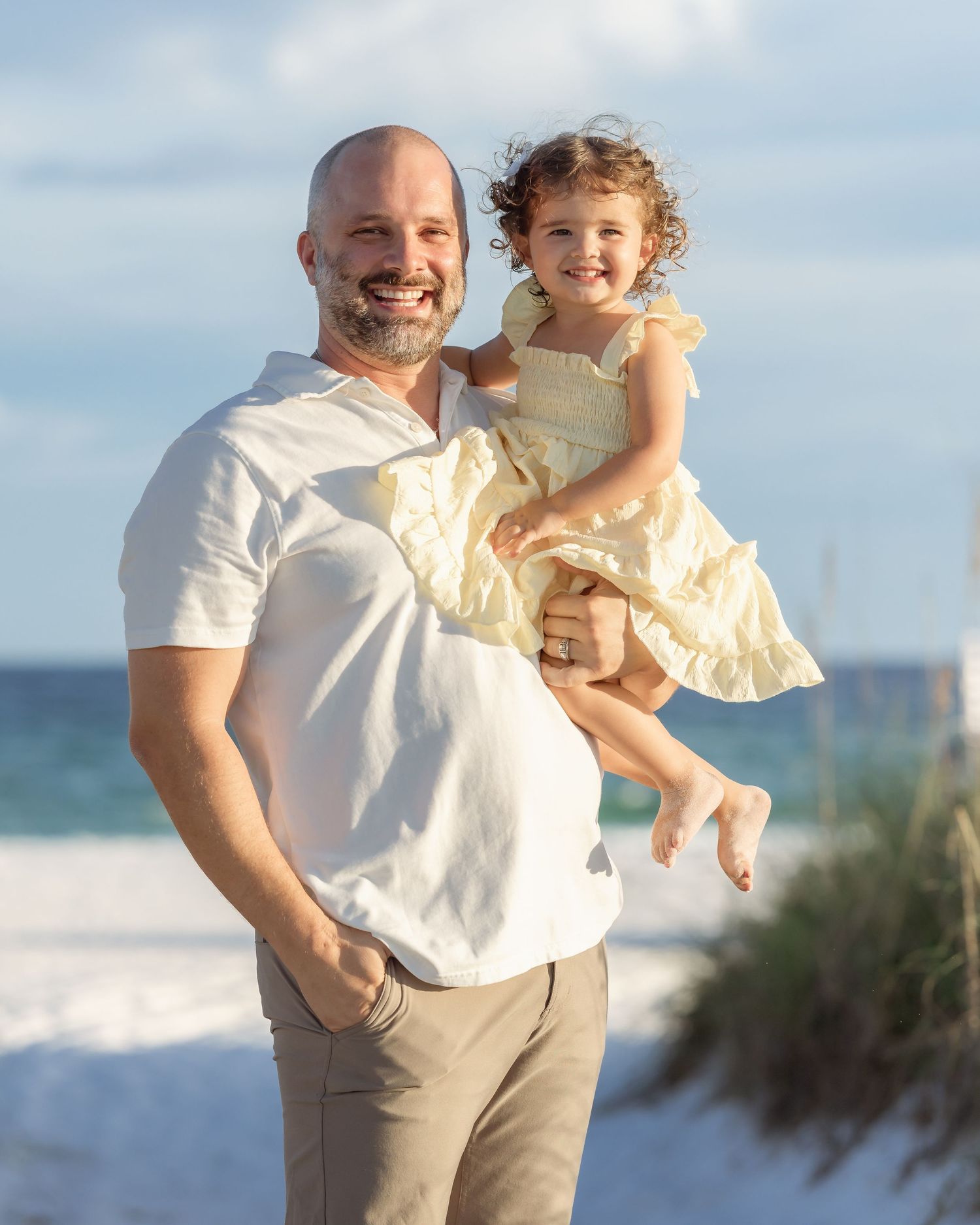 Smiling bald man holding a young girl in a yellow dress on a sunny beach.