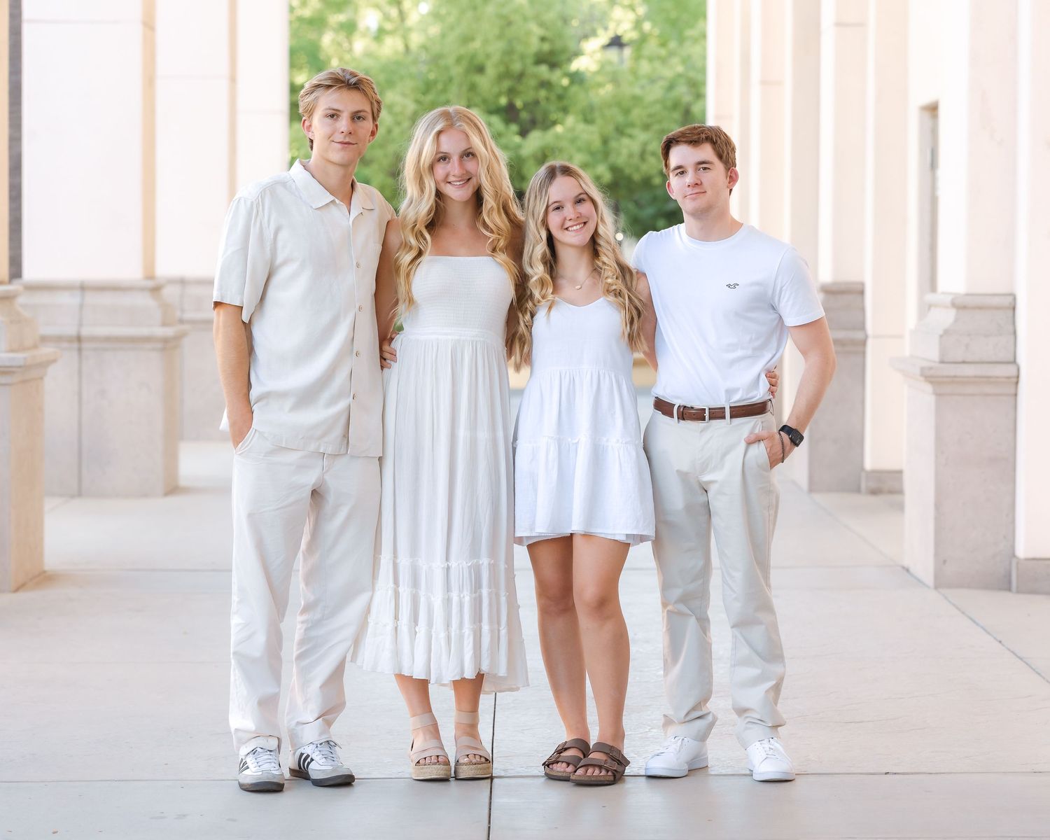 Four young adults in white and cream outfits posing together between columns in an elegant outdoor setting.