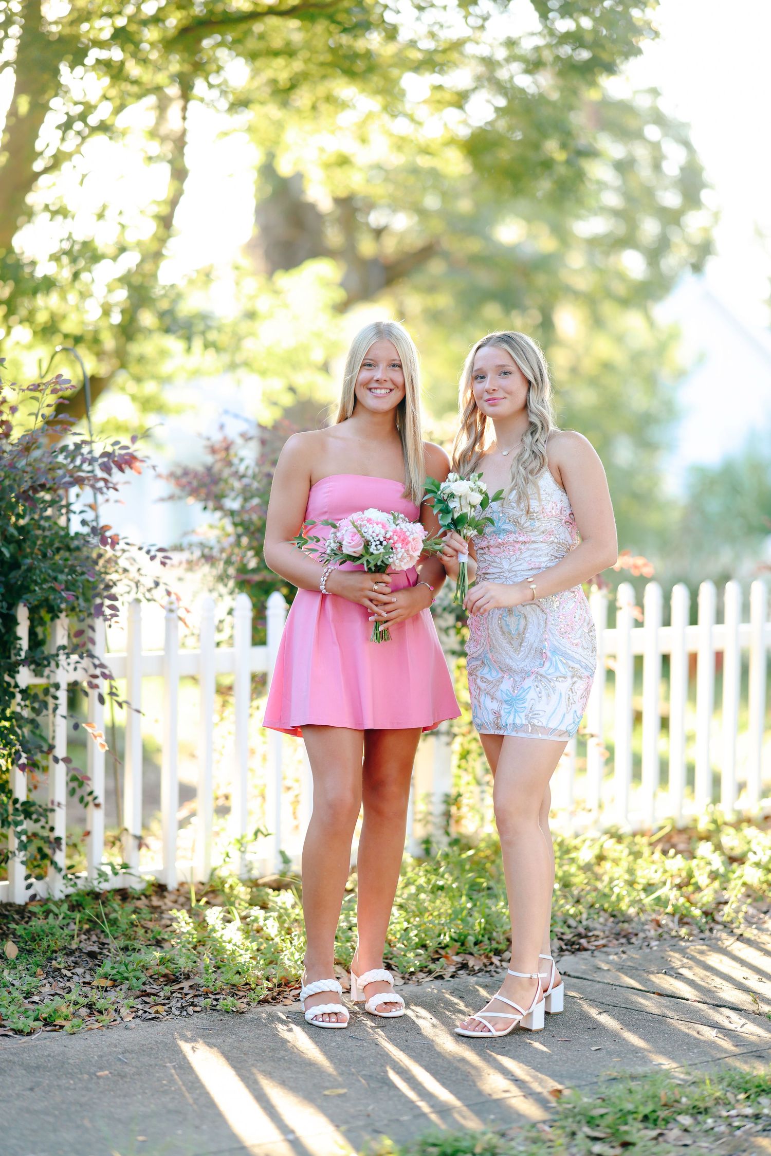 Two smiling teenage girls in formal dresses holding bouquets pose by a white picket fence outdoors.
