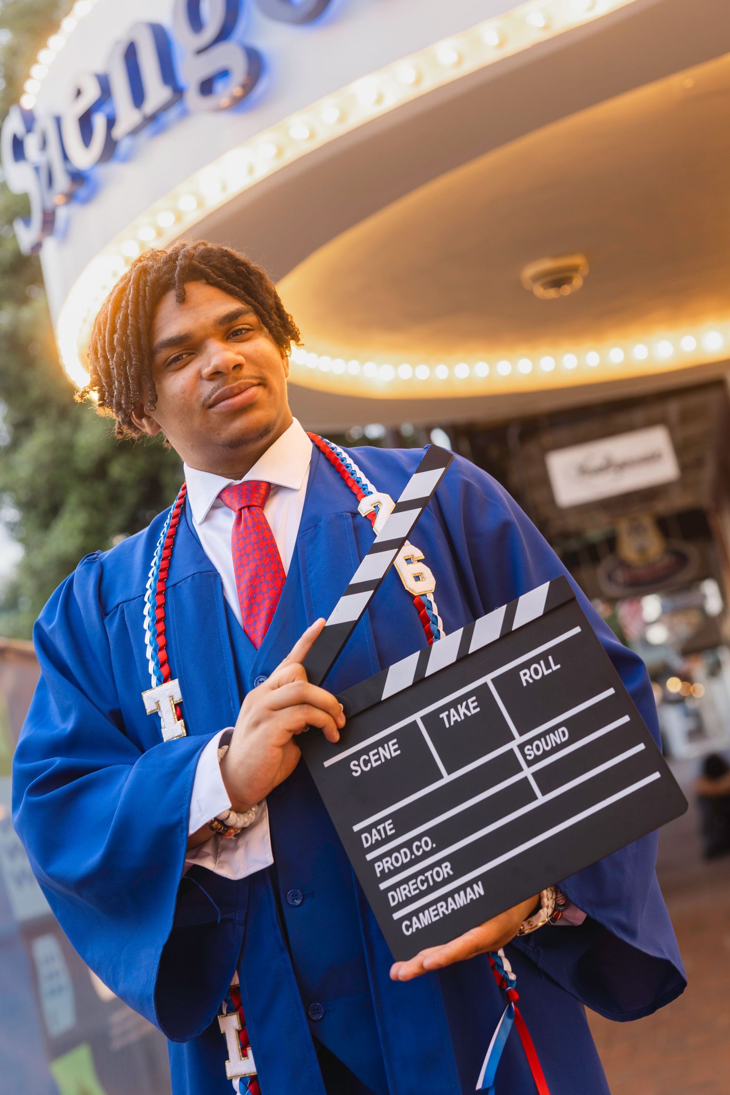 Graduate in blue cap and gown holds film clapperboard outside theater, wearing red tie and honor cords.