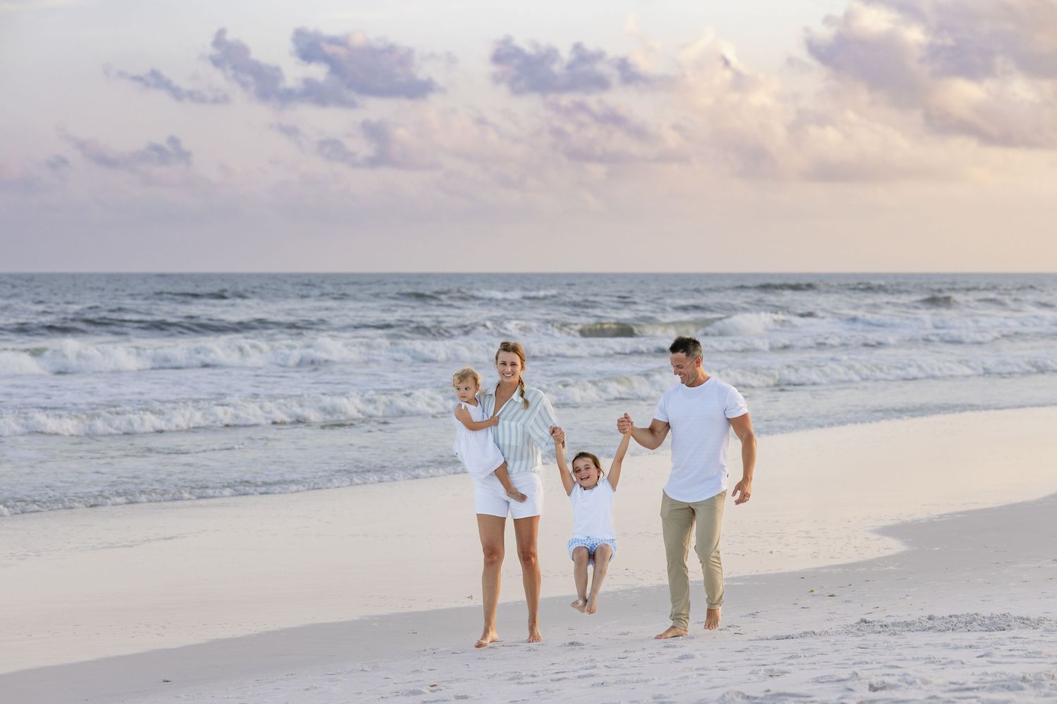 Family of four walking on a sandy beach at sunset, parents swinging children by hands near ocean waves.