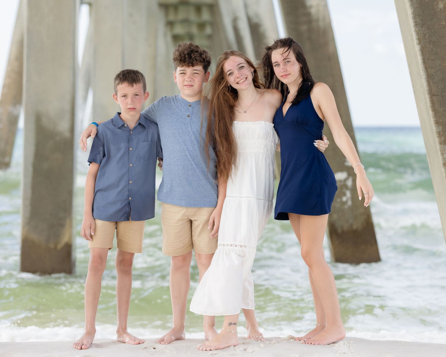 Four siblings posing barefoot on sandy beach near pier, two boys and two girls smiling in coordinated outfits.