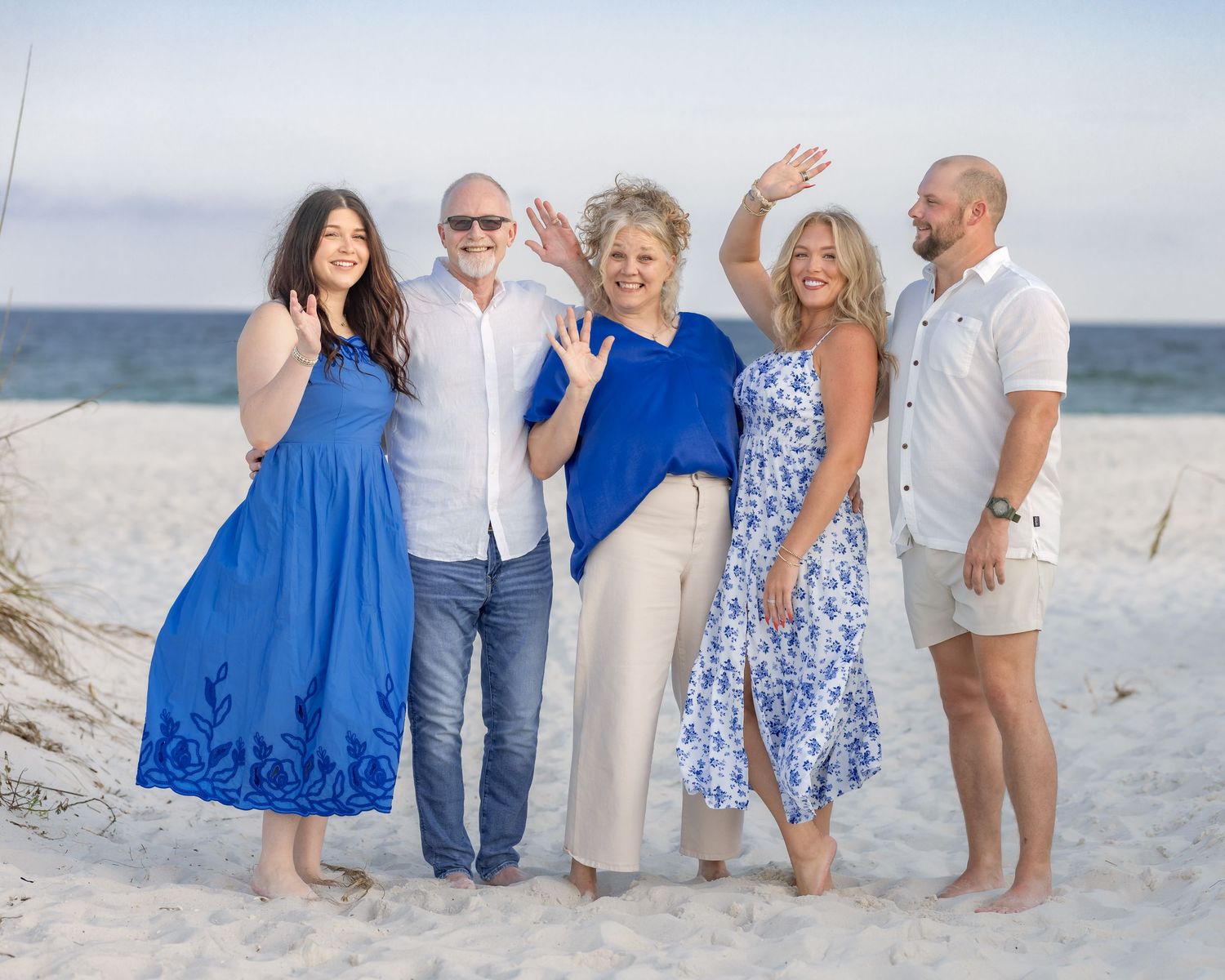 Happy family of five posing and waving on a sunny beach, dressed in blue and white outfits.