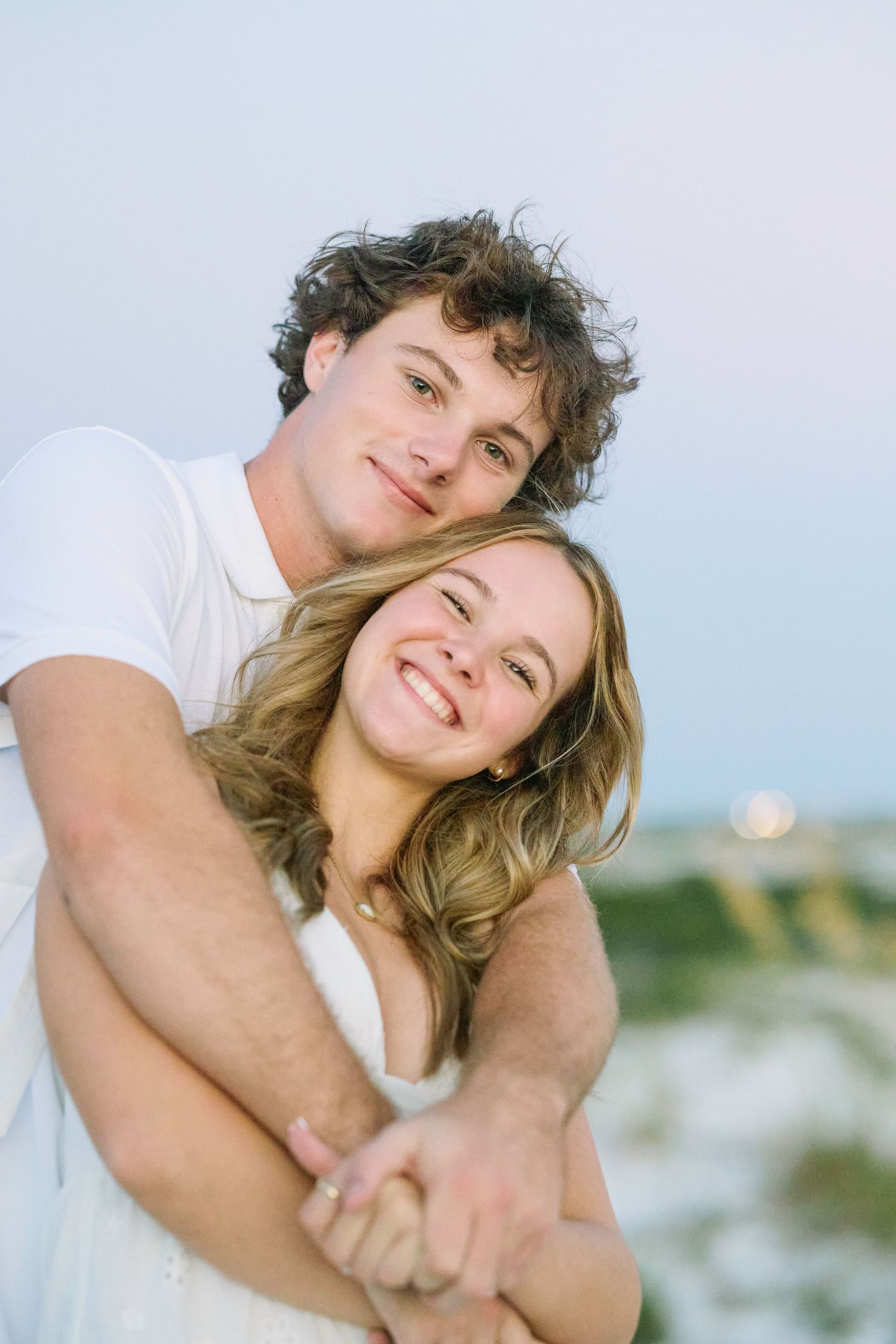 A couple shares a tender moment outdoors, dressed in white and smiling while embracing each other.