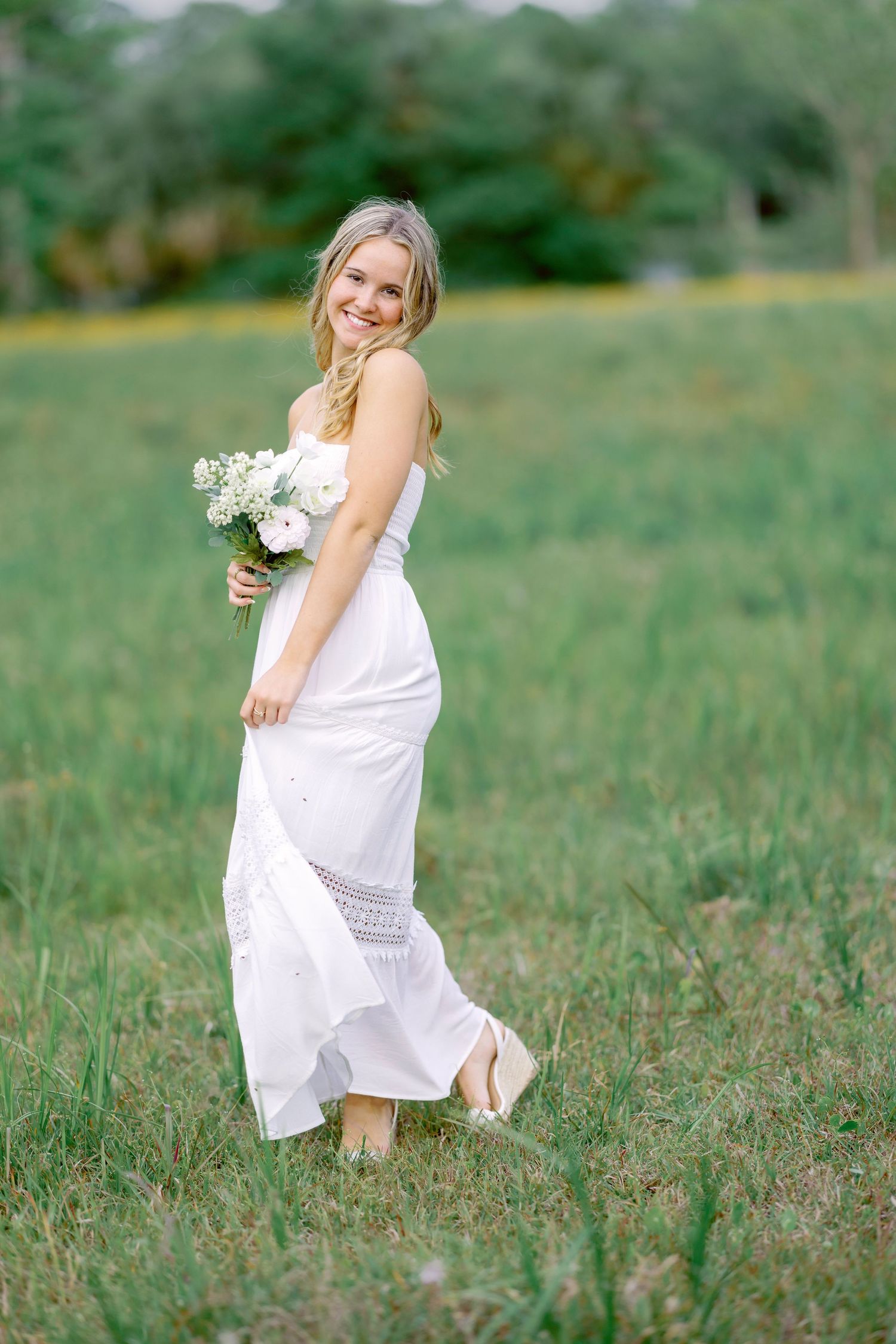 A person in a flowing white lace dress holds a bouquet while standing in a green summer meadow.