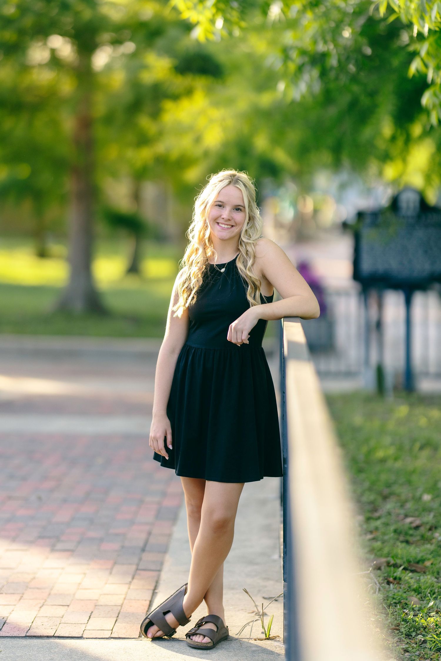 A person wearing a black summer dress poses playfully on a brick pathway surrounded by lush green trees in a park setting.