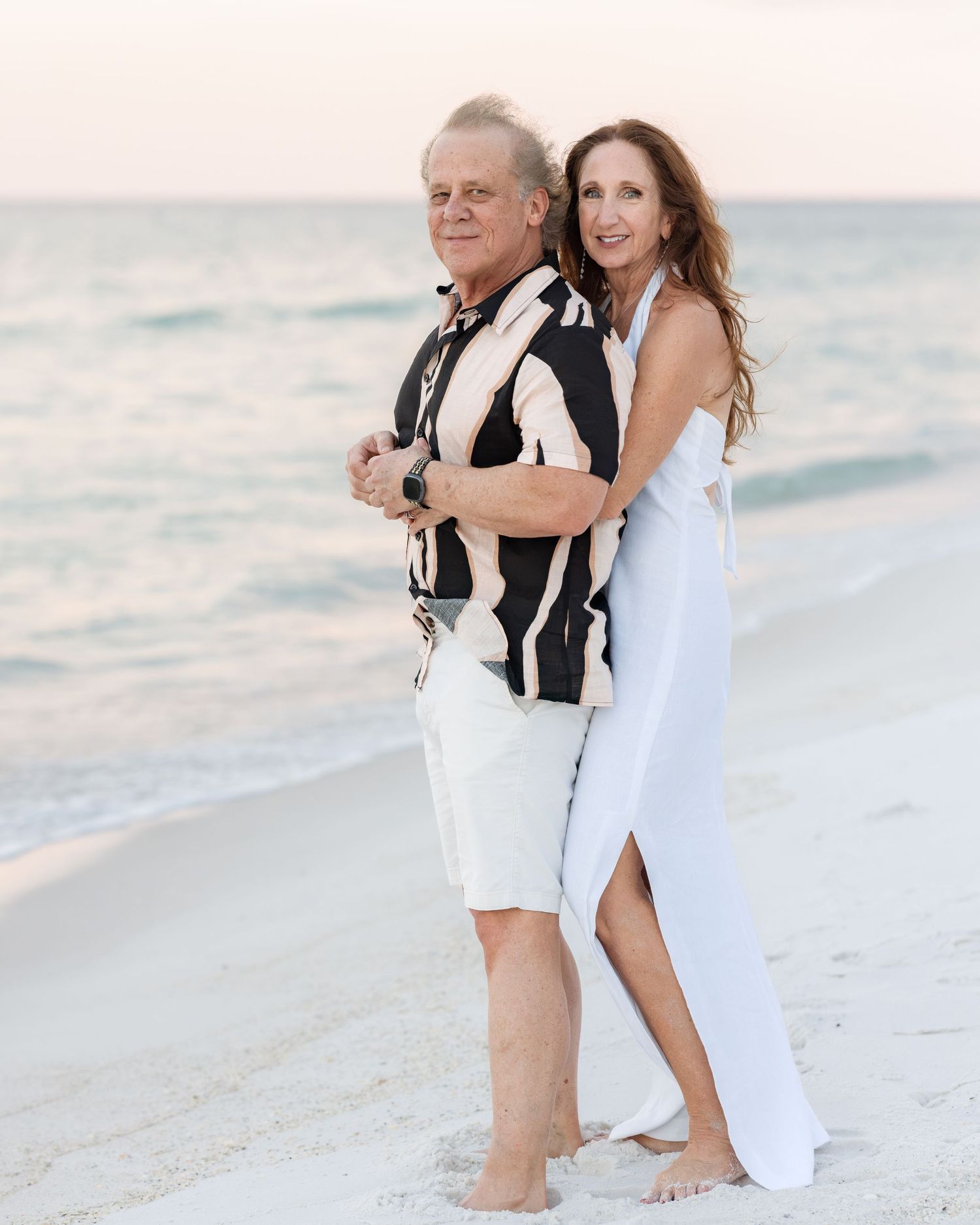 Elegant older couple embracing on white sand beach at sunset in coordinated outfits.