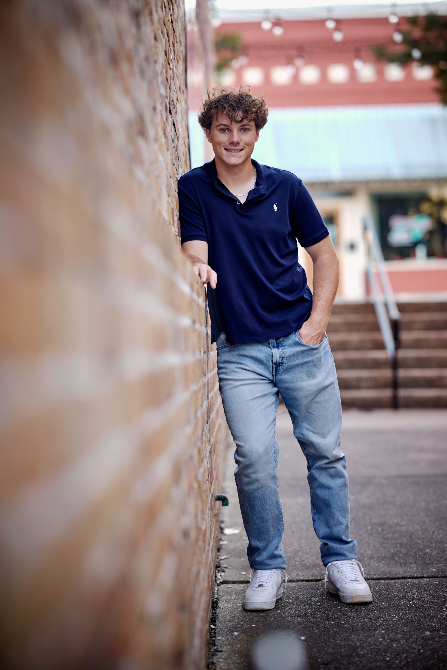 Young man in navy polo and jeans leaning against brick wall, smiling confidently in an outdoor urban setting.