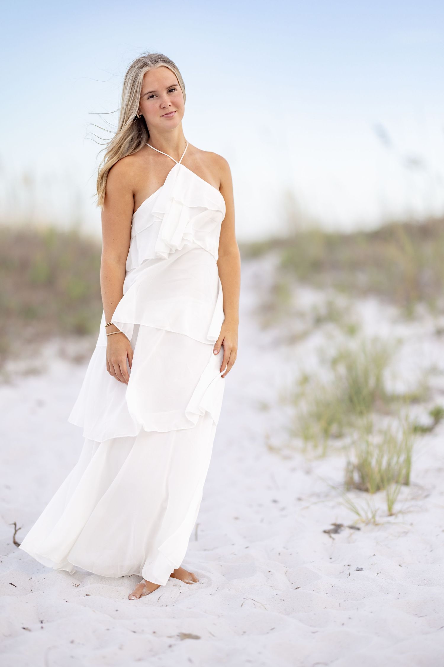 Young woman in white ruffled halter maxi dress standing barefoot on white sand beach dunes.