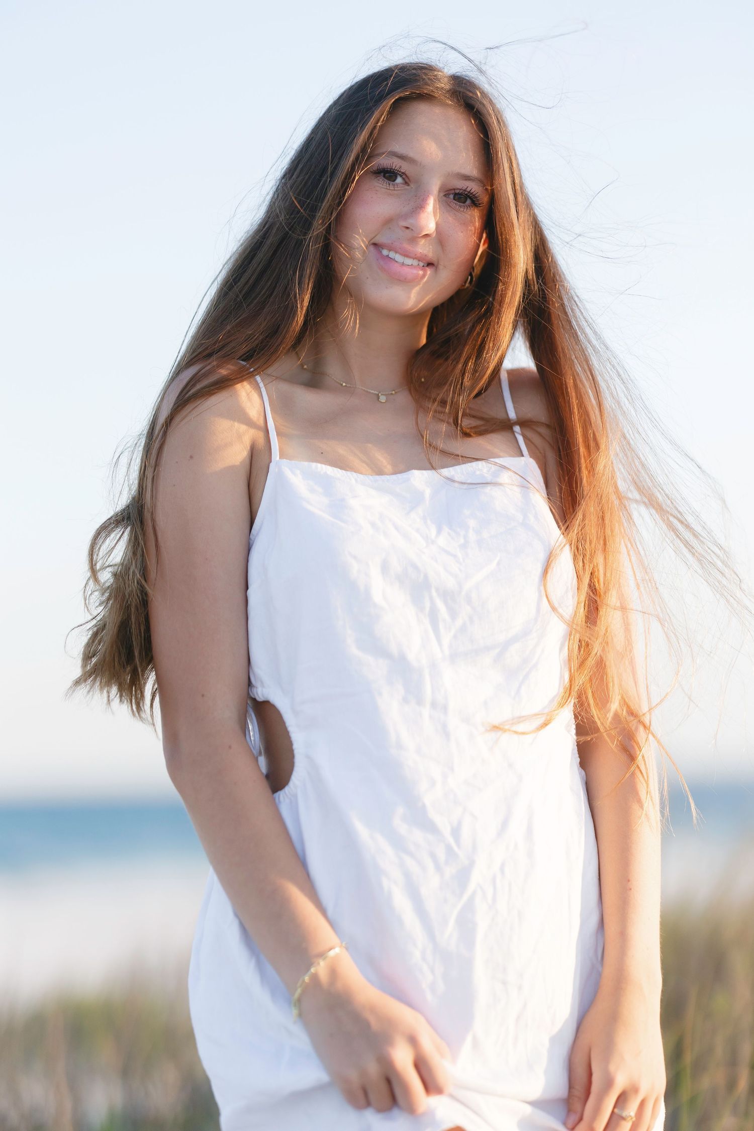 Woman in flowing white dress with windswept hair poses on beach at sunset.